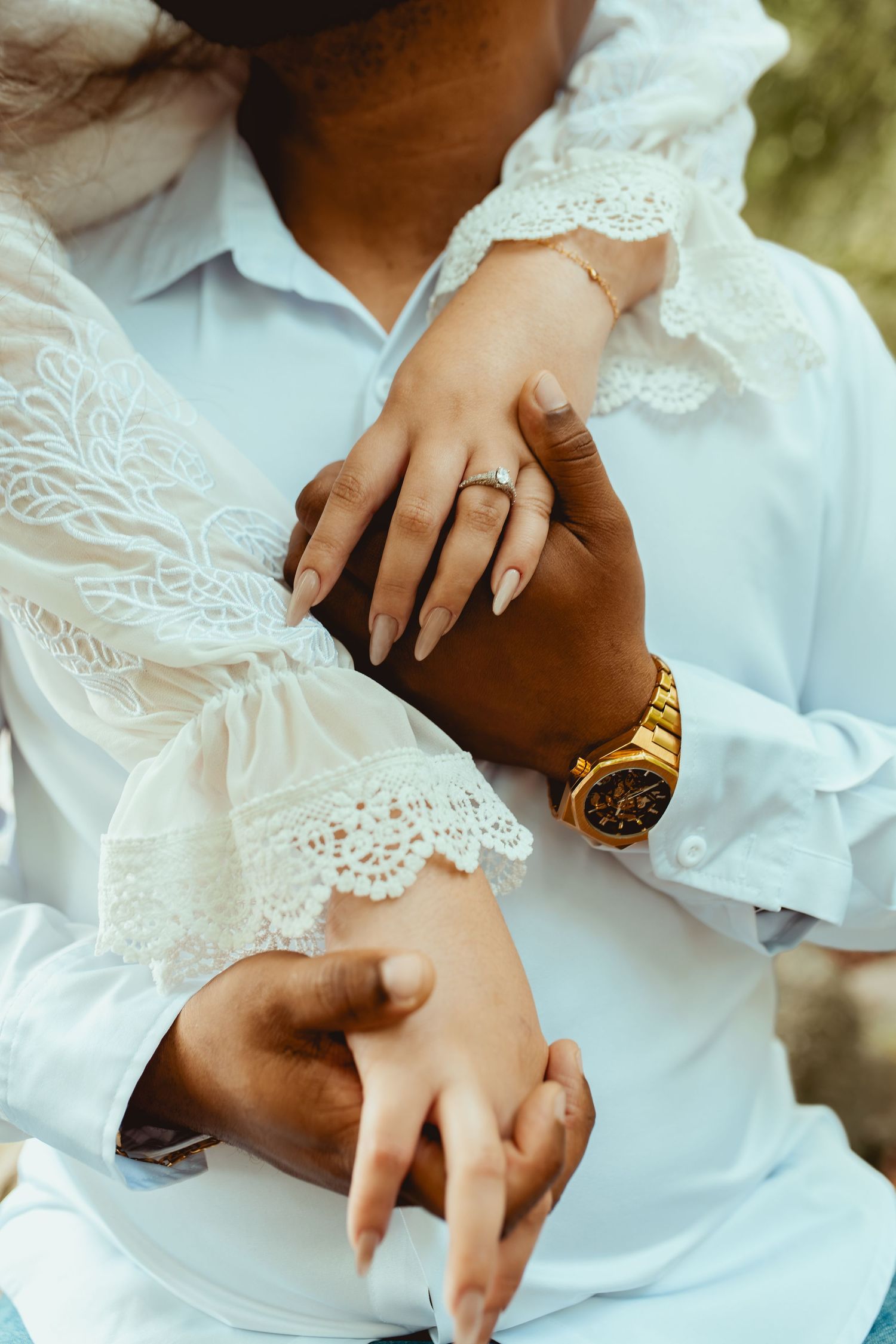 Interracial couple embracing, woman in white lace dress with ring, man in blue shirt wearing gold watch.