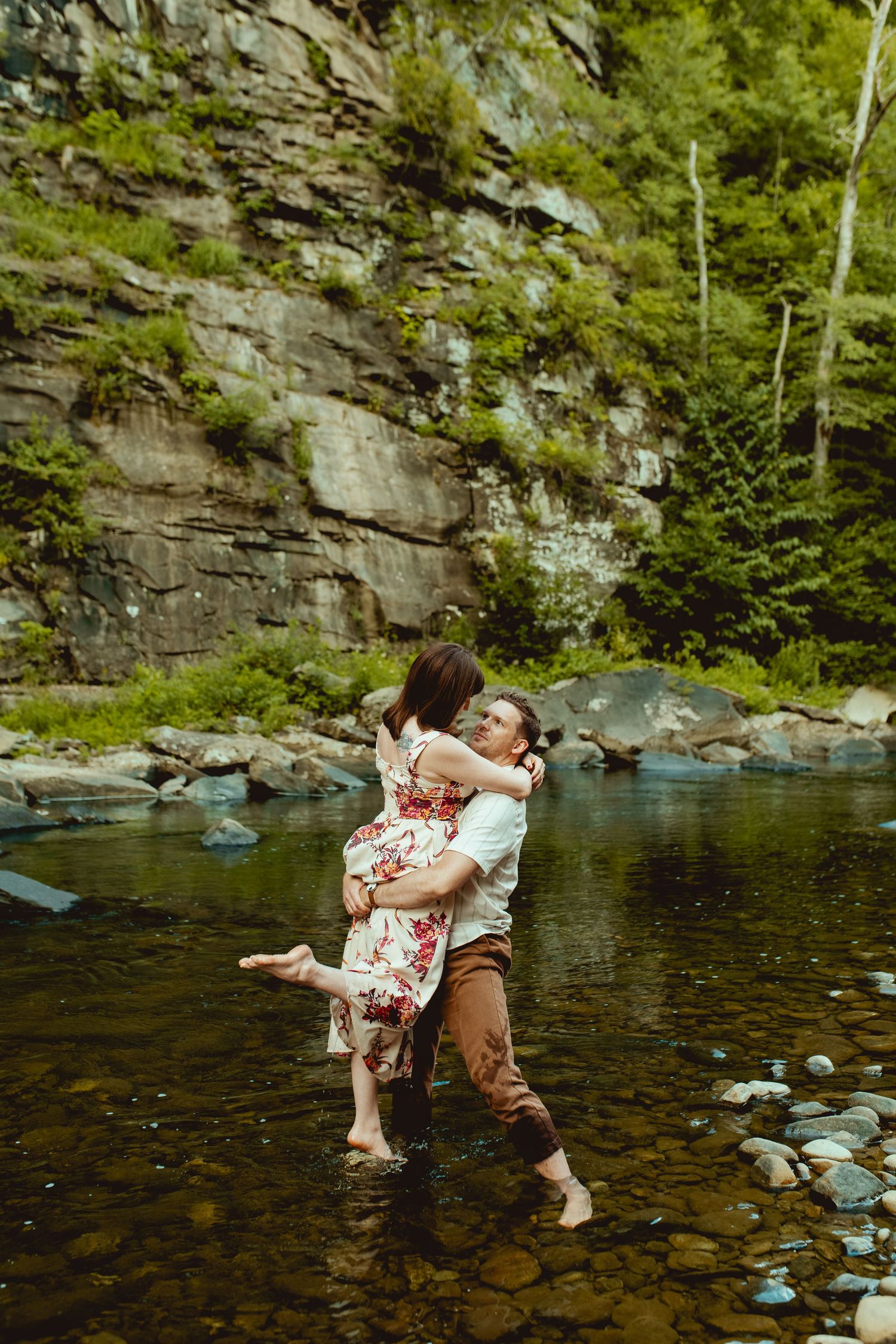 Couple embracing in a rocky stream surrounded by lush greenery and a stone cliff backdrop.