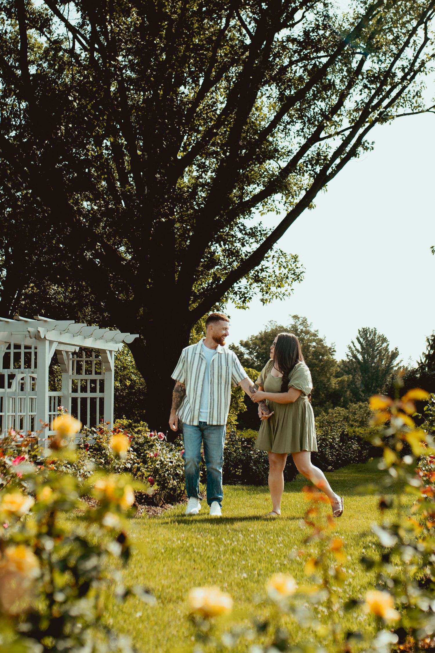 Couple holding hands and dancing in a lush garden with yellow roses and a white pergola on a sunny day.