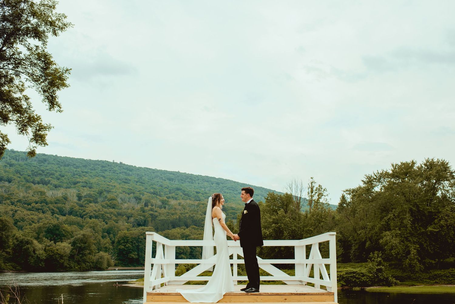 Couple standing on white wooden bridge overlooking mountain landscape on wedding day.