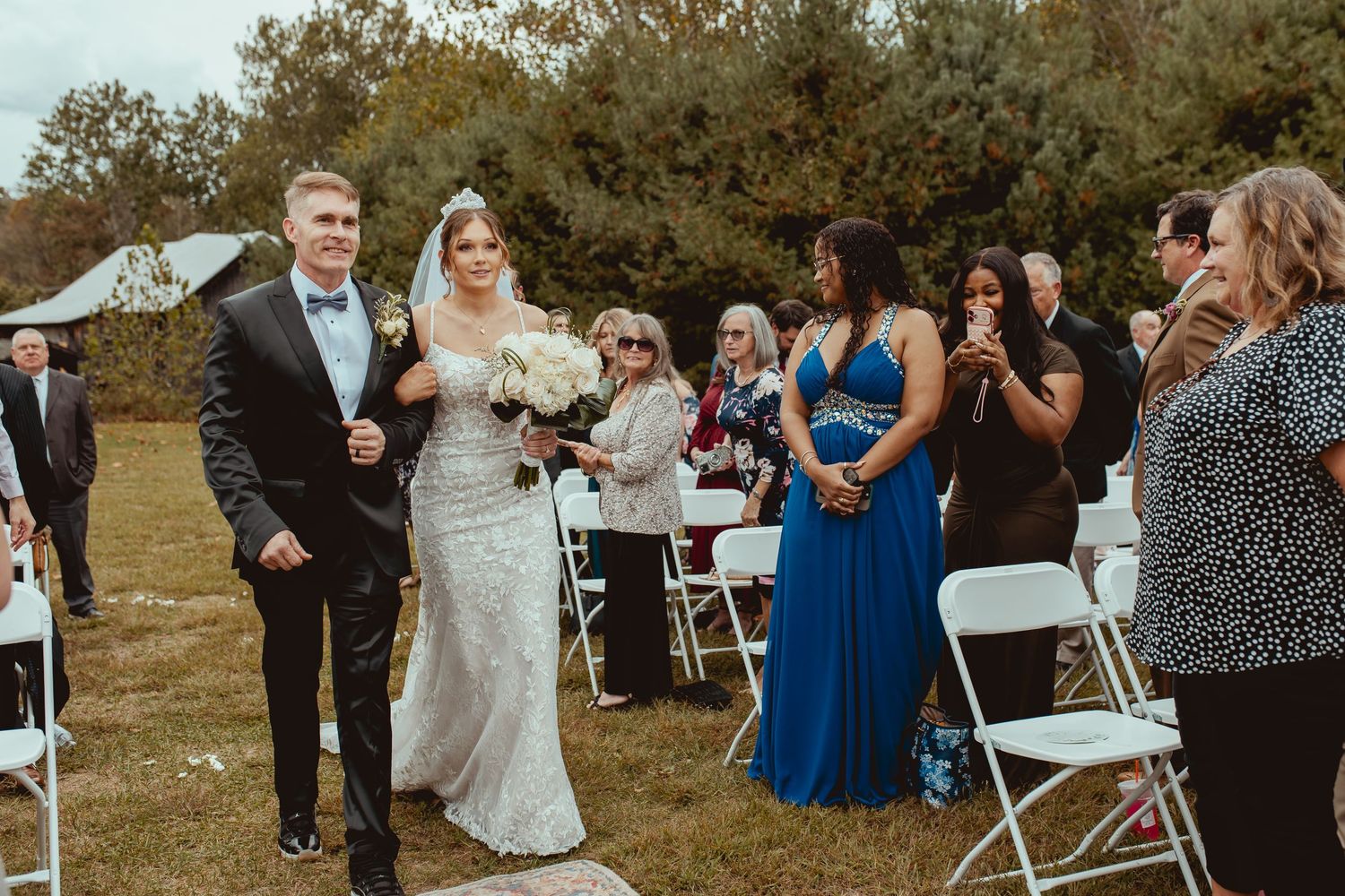 An outdoor wedding ceremony with guests seated in white chairs against an autumn backdrop.