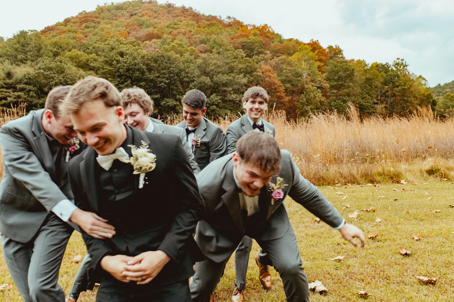 Groomsmen in grey suits laughing and joking around in a field during wedding photos.