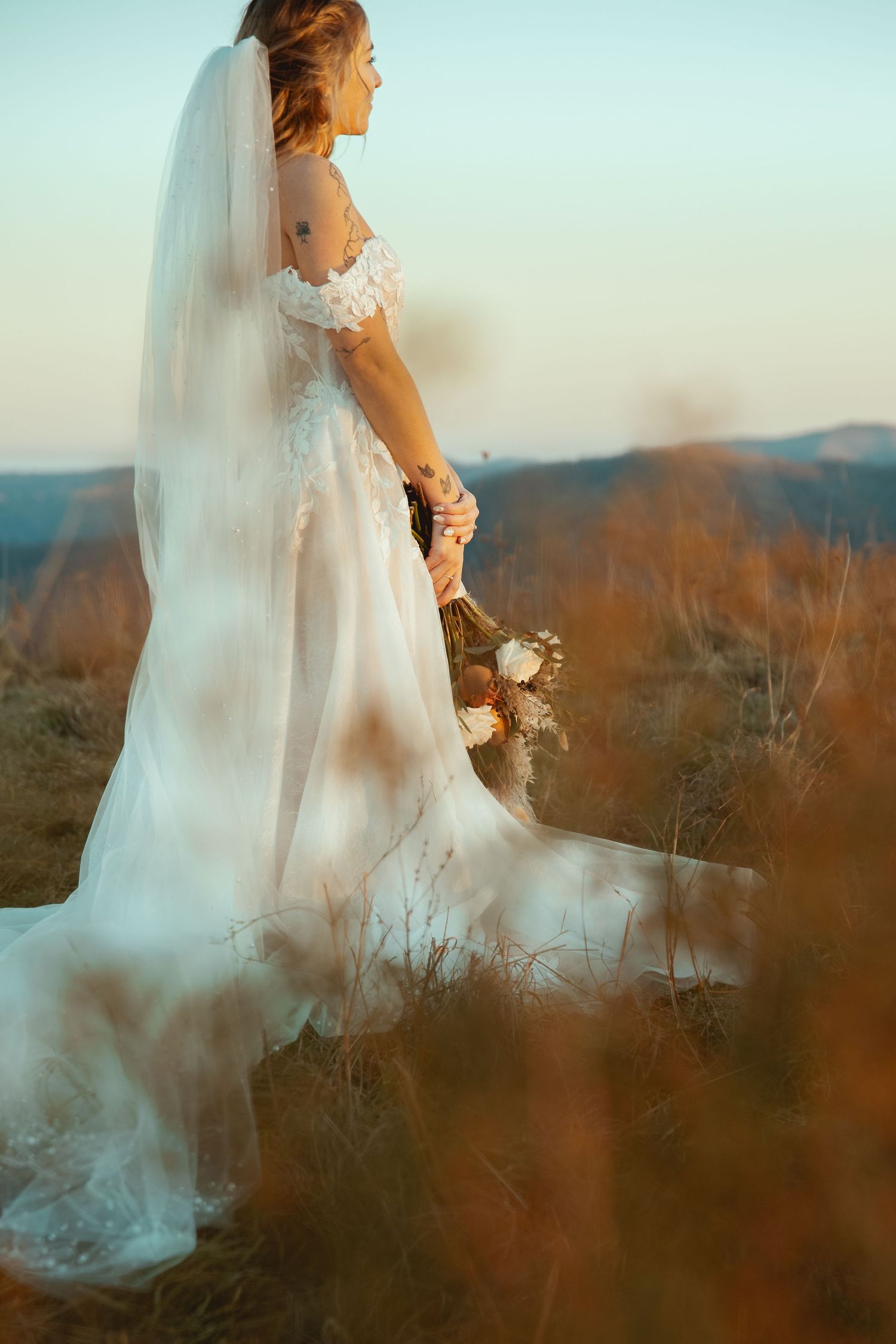 An ethereal bridal portrait with flowing dress and veil against a sunset mountain landscape.
