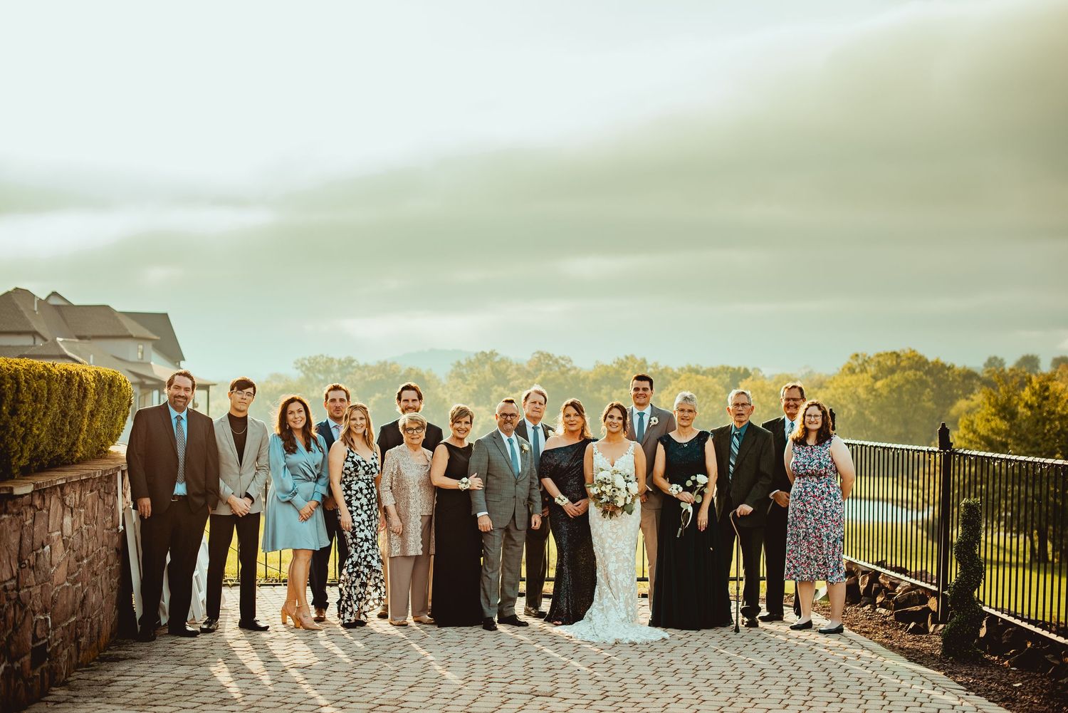 Wedding party poses together on stone terrace overlooking scenic countryside on cloudy autumn day.