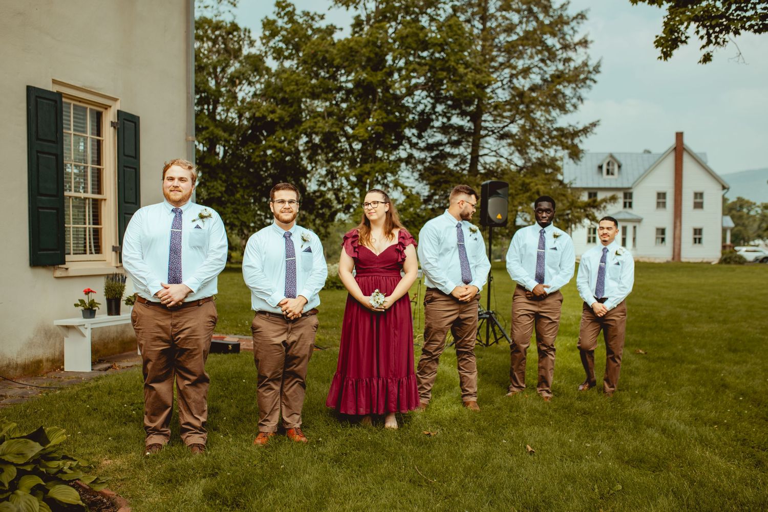 A wedding party poses on a lawn in front of a white colonial building with brown pants and blue shirts.