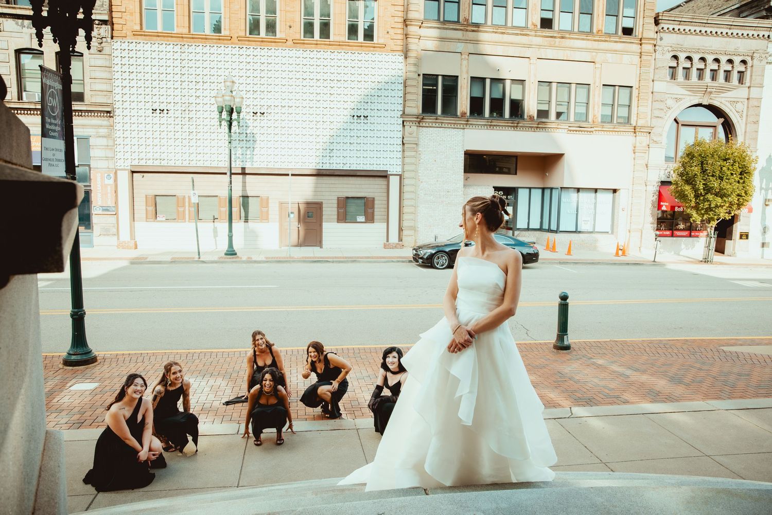 A wedding party poses for photos on a stone patio outside an ornate building with columns and archways.