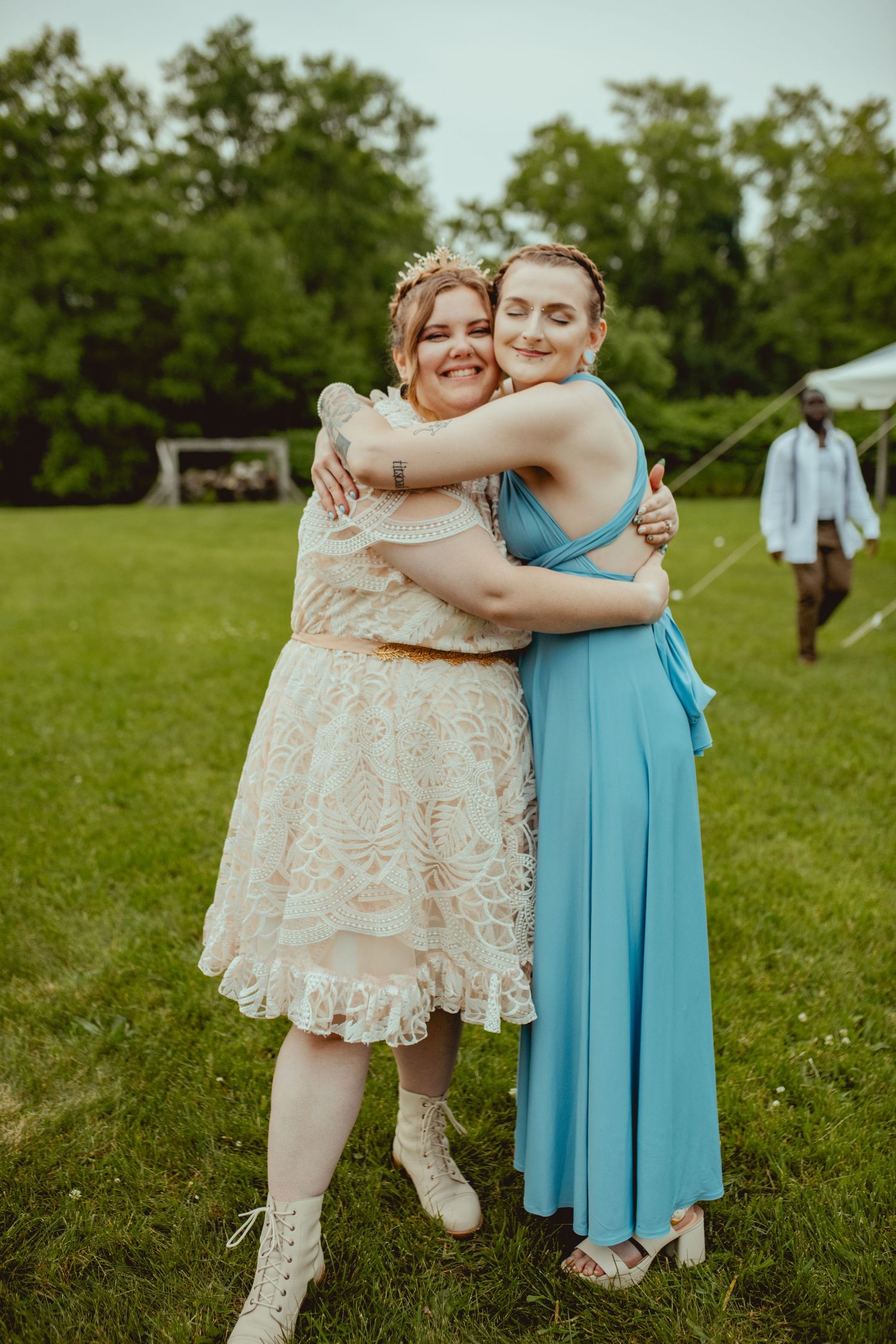 Two people in light colored dresses embrace while standing on green grass during a summer gathering.
