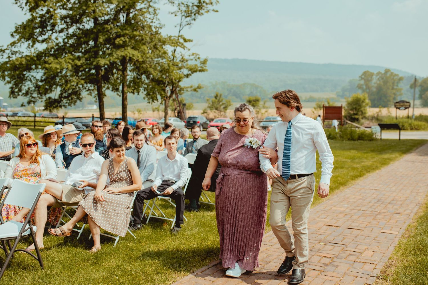 Guests enjoy a casual outdoor celebration with tables and chairs set up along a tree-lined path on a sunny day.