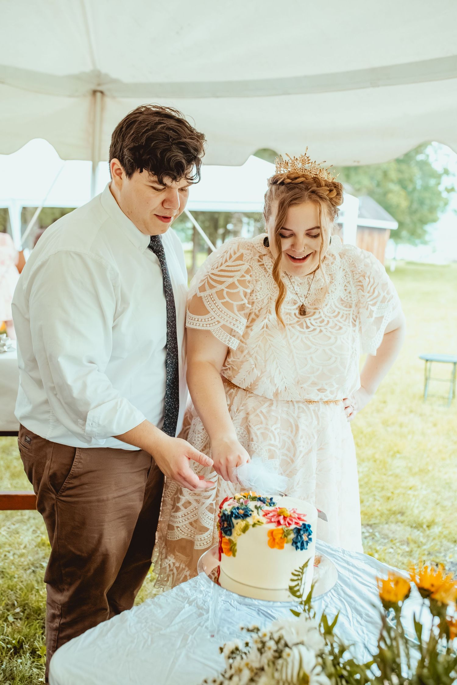 Newlyweds cut their white wedding cake together at an outdoor reception with colorful floral decorations.