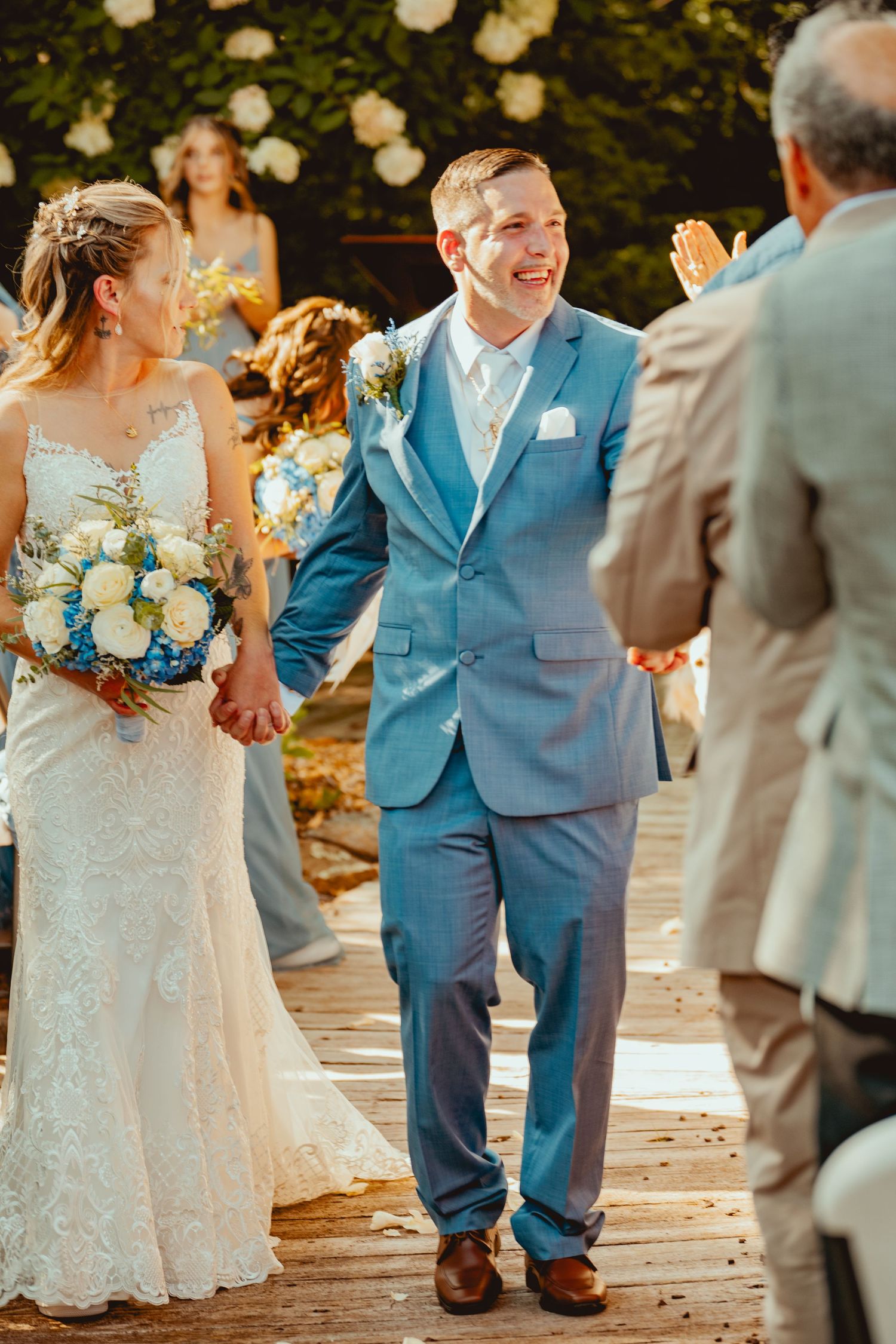 Wedding party in blue and gray suits gathered outdoors with floral bouquets.