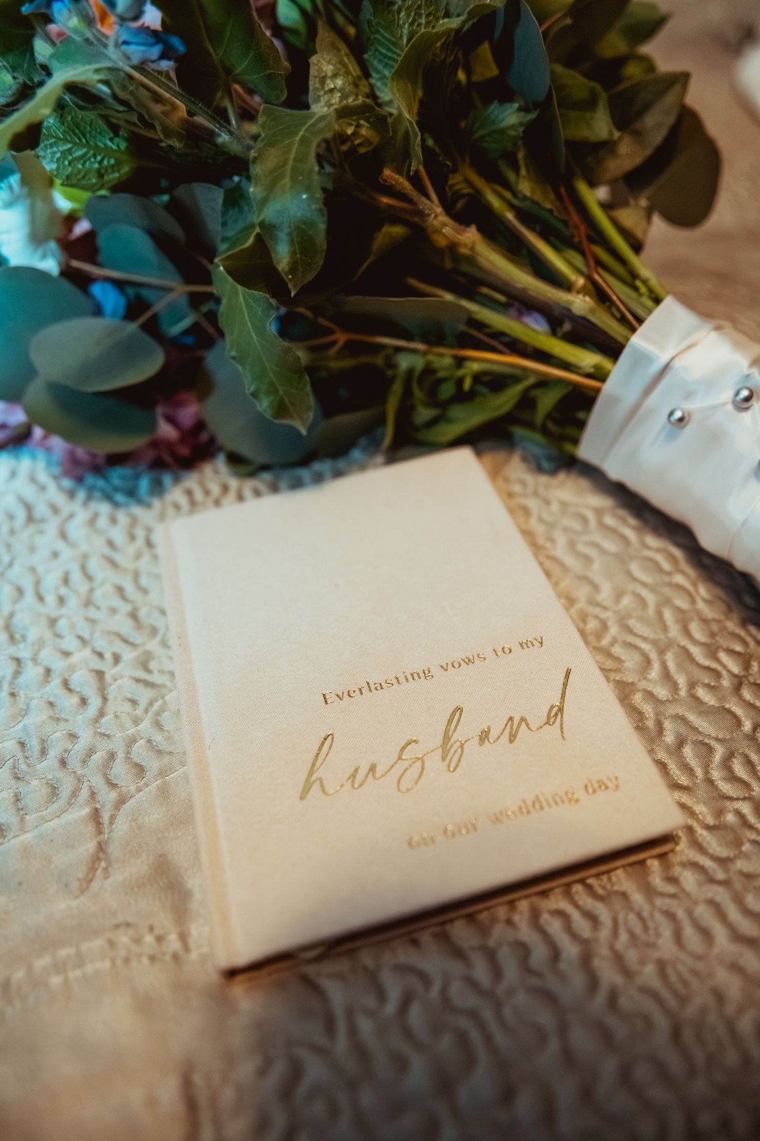 A cream vow book engraved with everlasting vows to my husband rests beside a bridal bouquet of greenery.