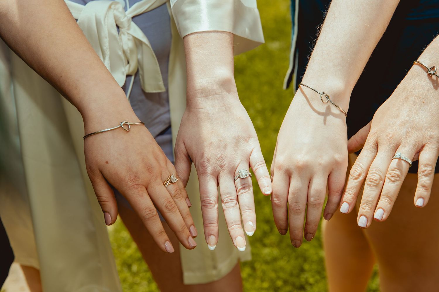 Three women displaying delicate gold bracelets and rings on their outstretched hands outdoors.