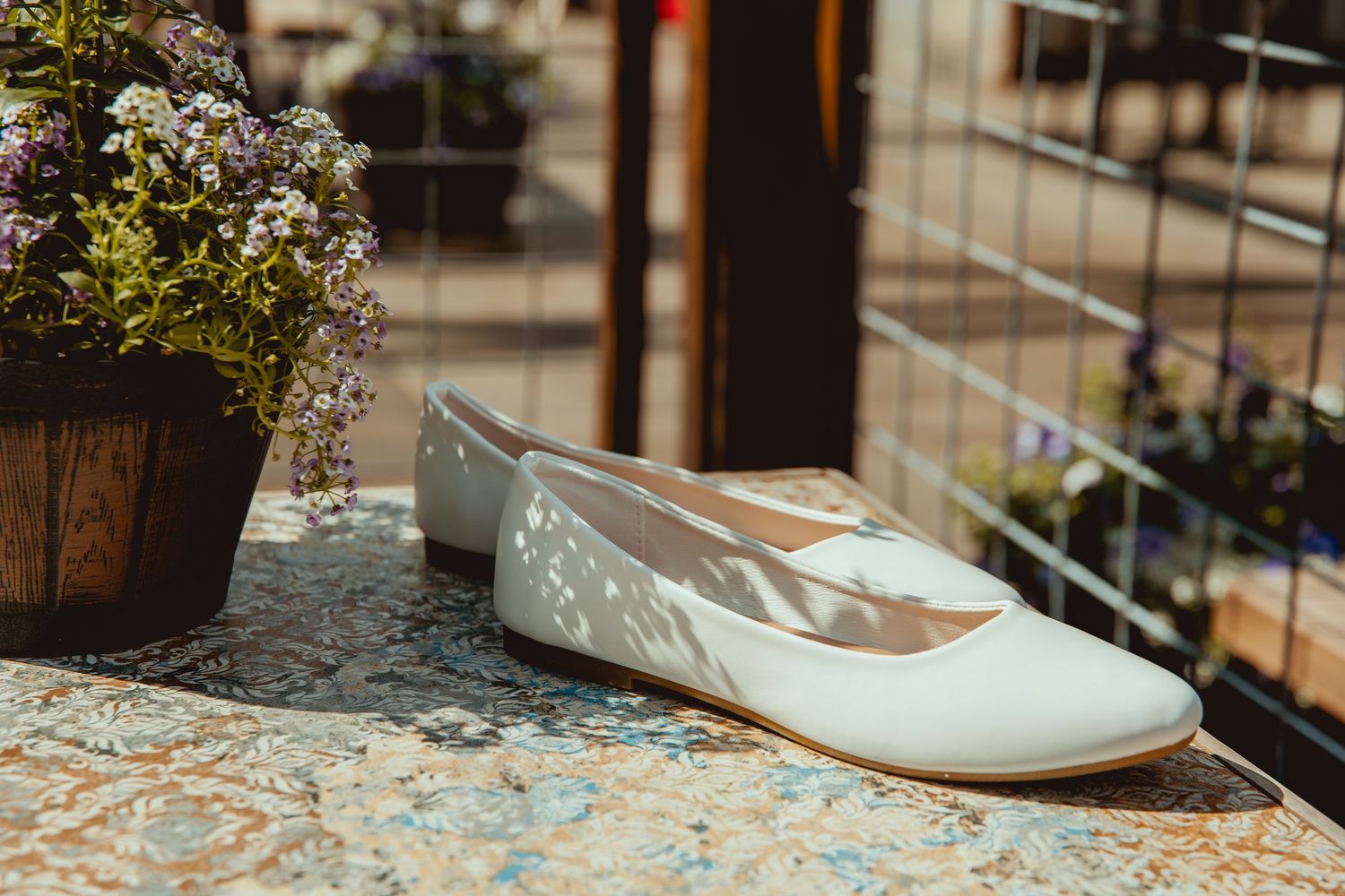 White leather ballet flat shoe displayed on a marble surface beside a flower pot in a sunlit outdoor setting.