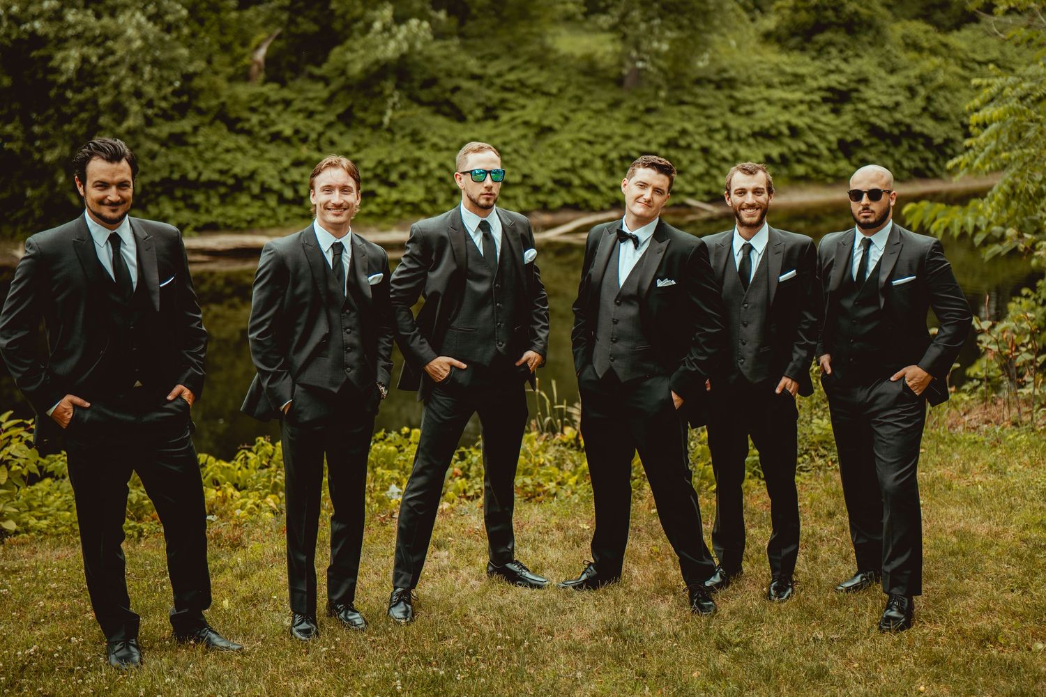 Line of groomsmen in black suits posing on grass for wedding photos.
