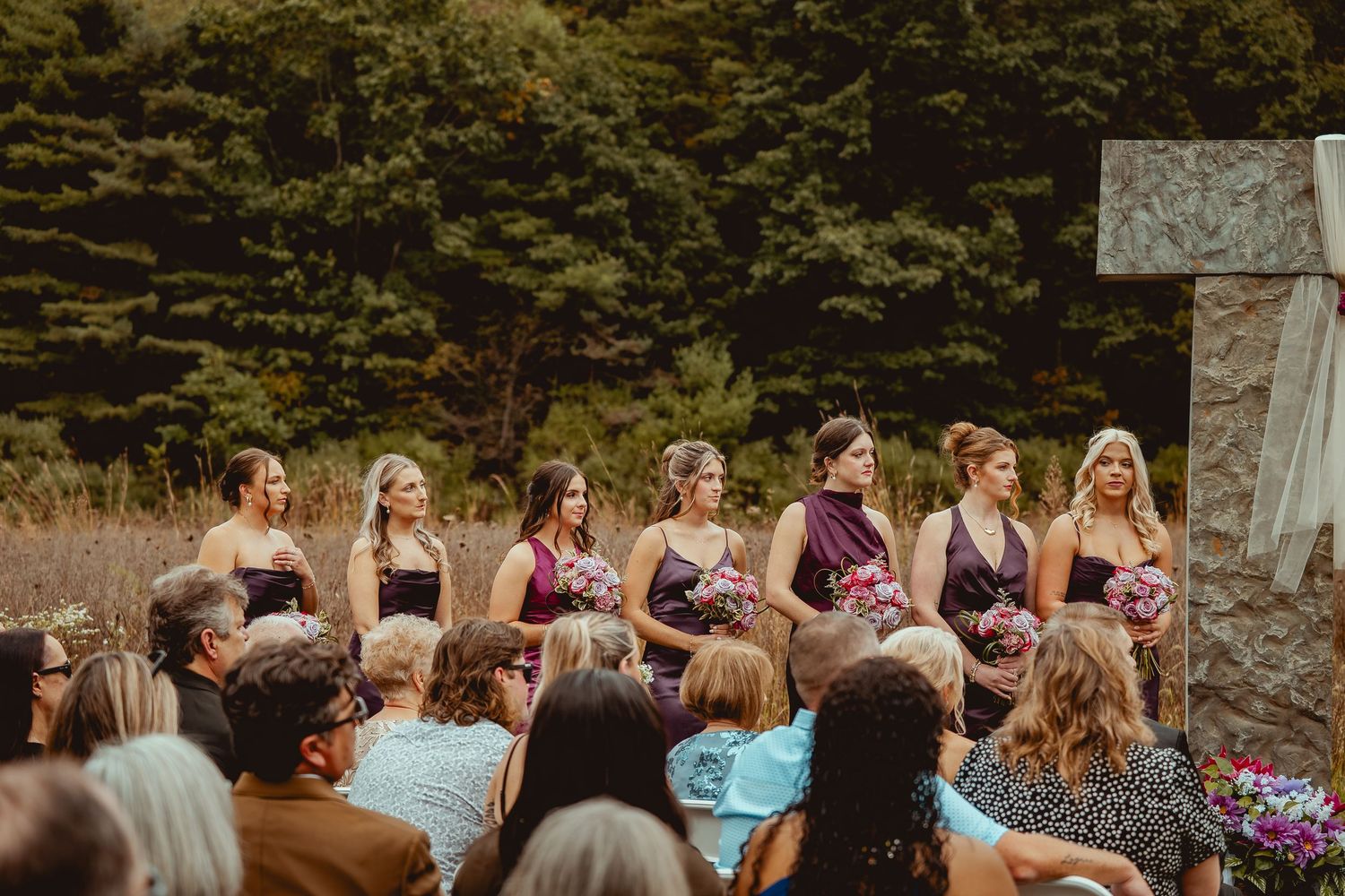 A group in burgundy and navy dresses stands in a line at an outdoor wedding ceremony with guests seated below.