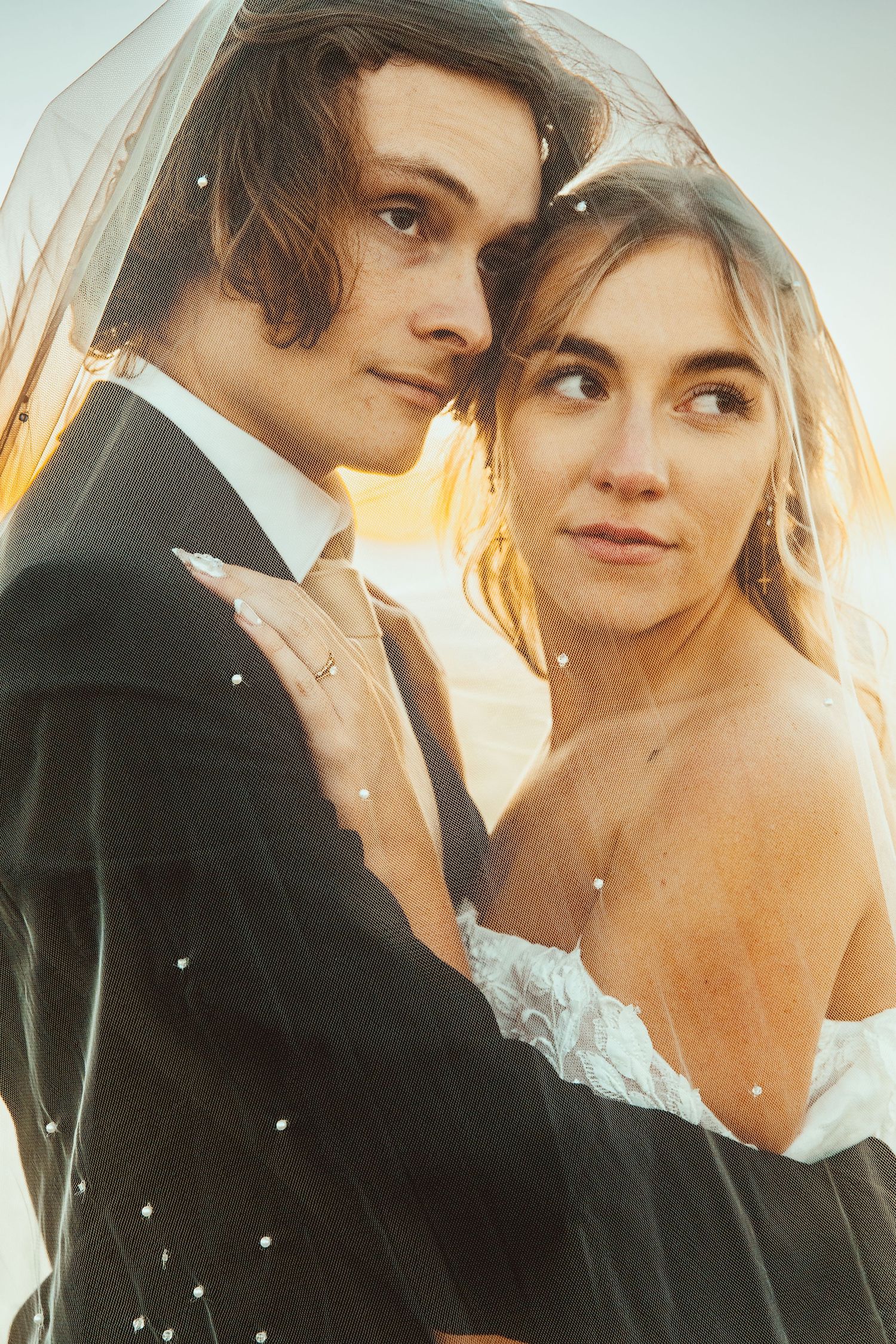 A romantic wedding portrait captures a couple beneath a veil with golden sunset light glowing behind them.