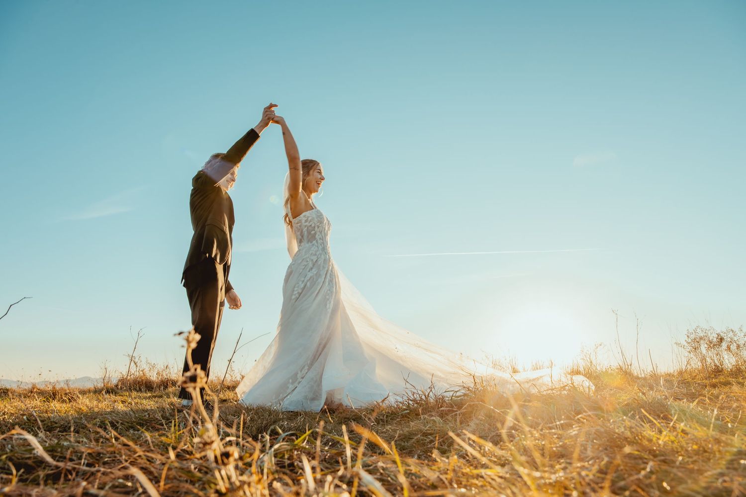 Newlywed couple dancing together in a golden field at sunset as her white wedding dress flows in the breeze.
