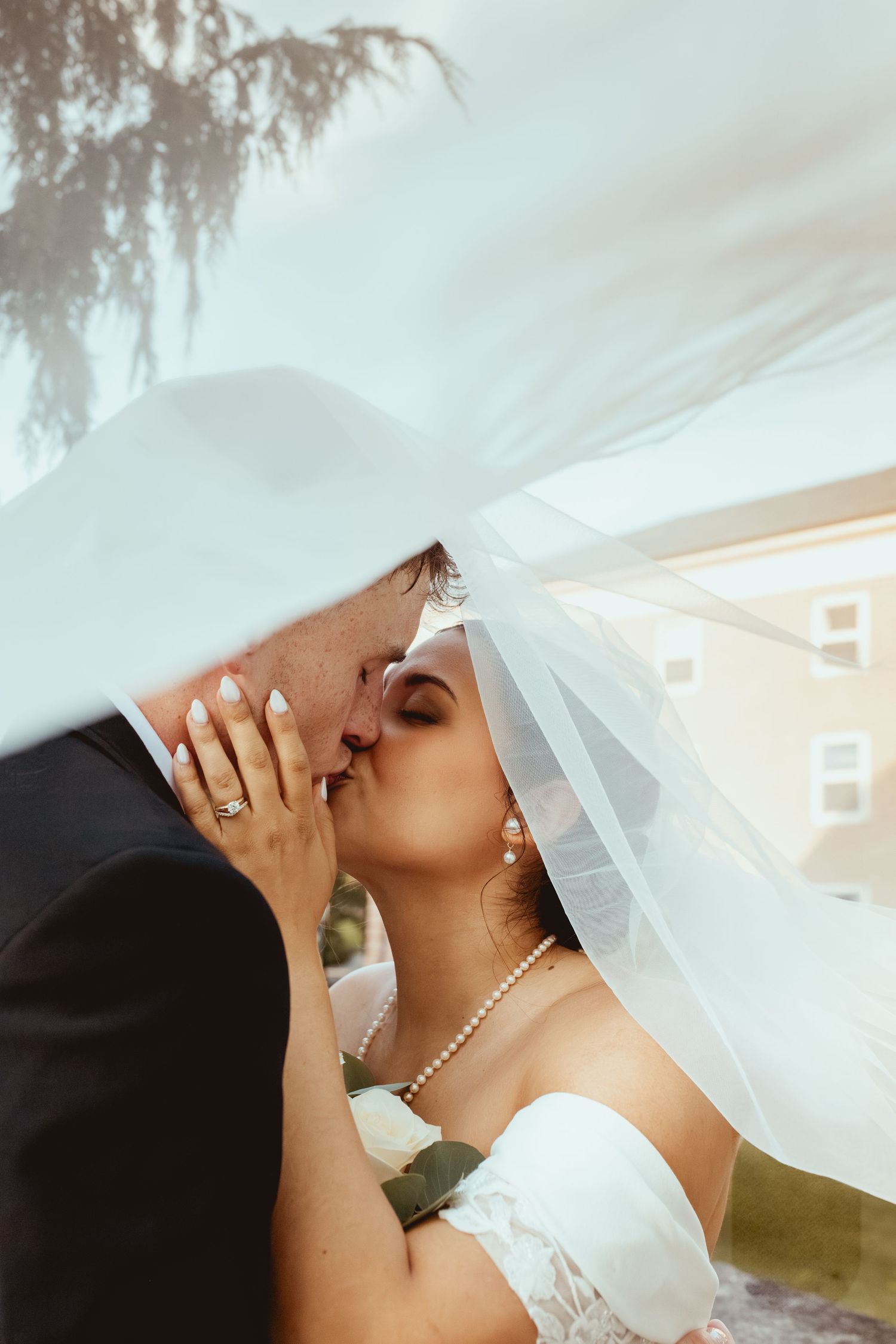 A romantic wedding kiss captured under a flowing white veil with pearl necklace details.