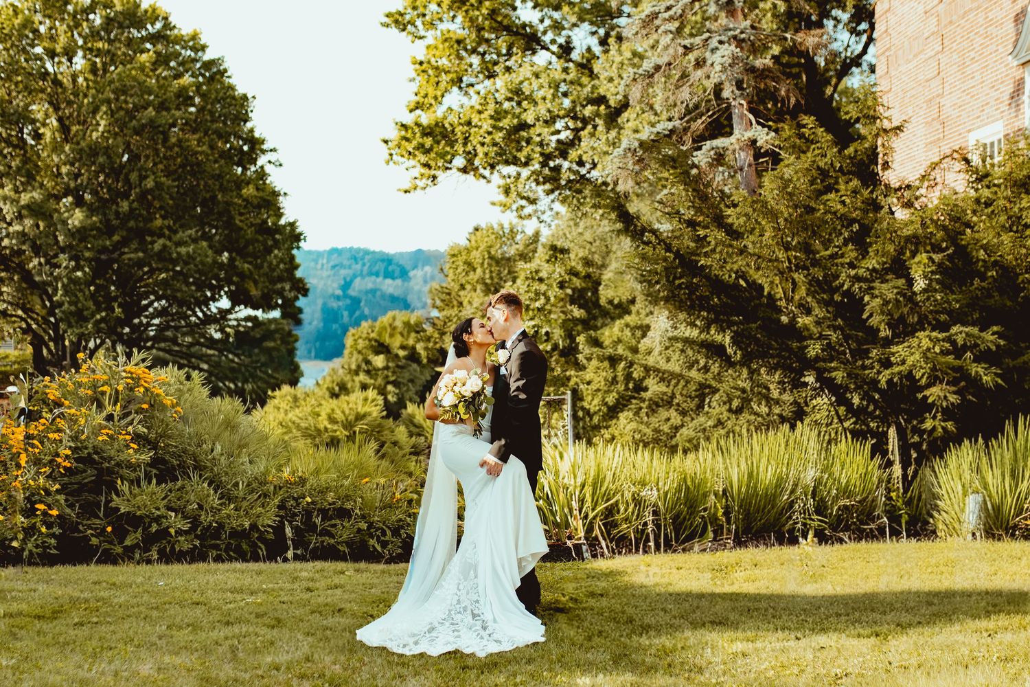 A romantic couple in light blue attire poses together in a sunlit garden setting with lush greenery and trees.