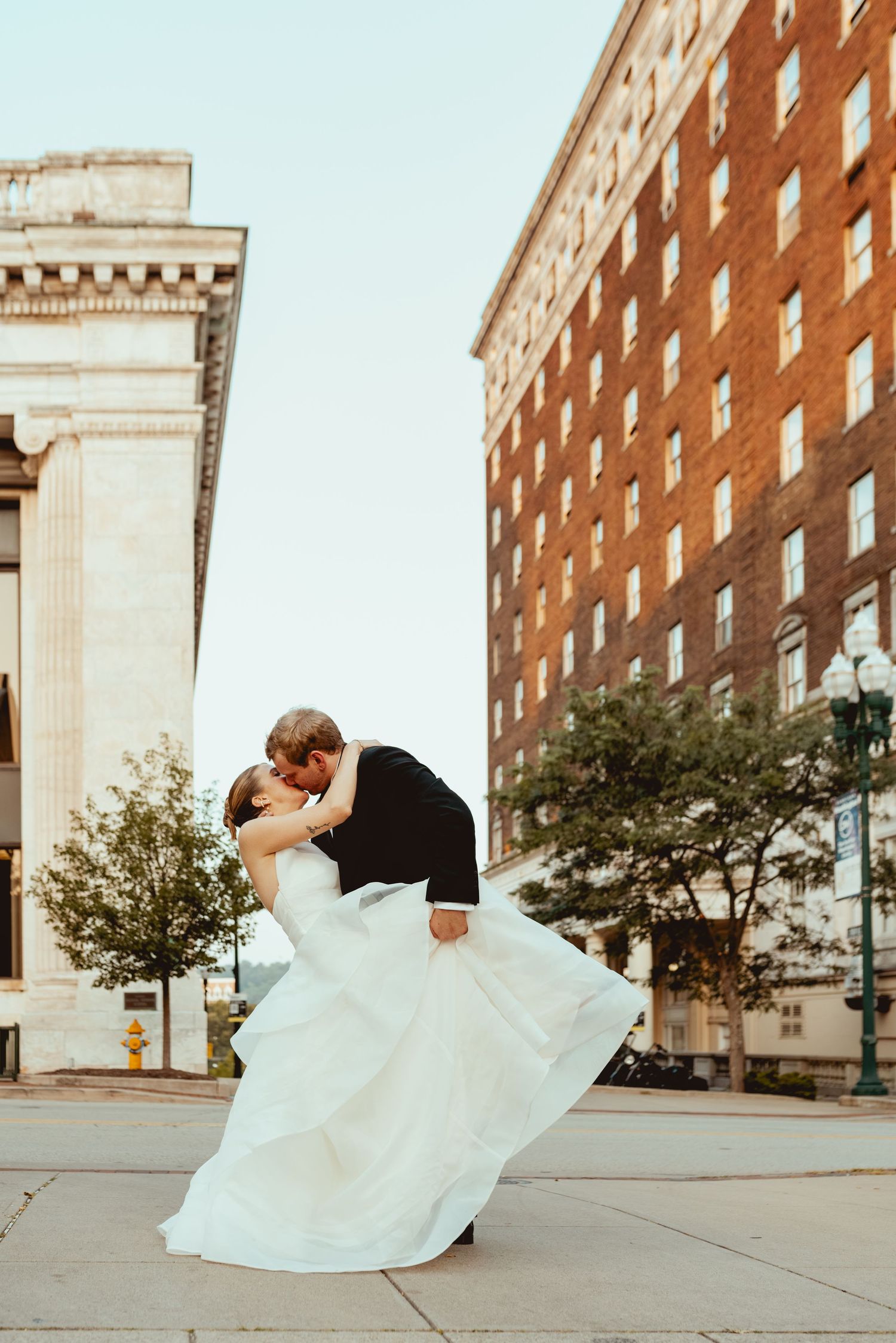 Romantic wedding couple dancing and dipping on city sidewalk near brick building.