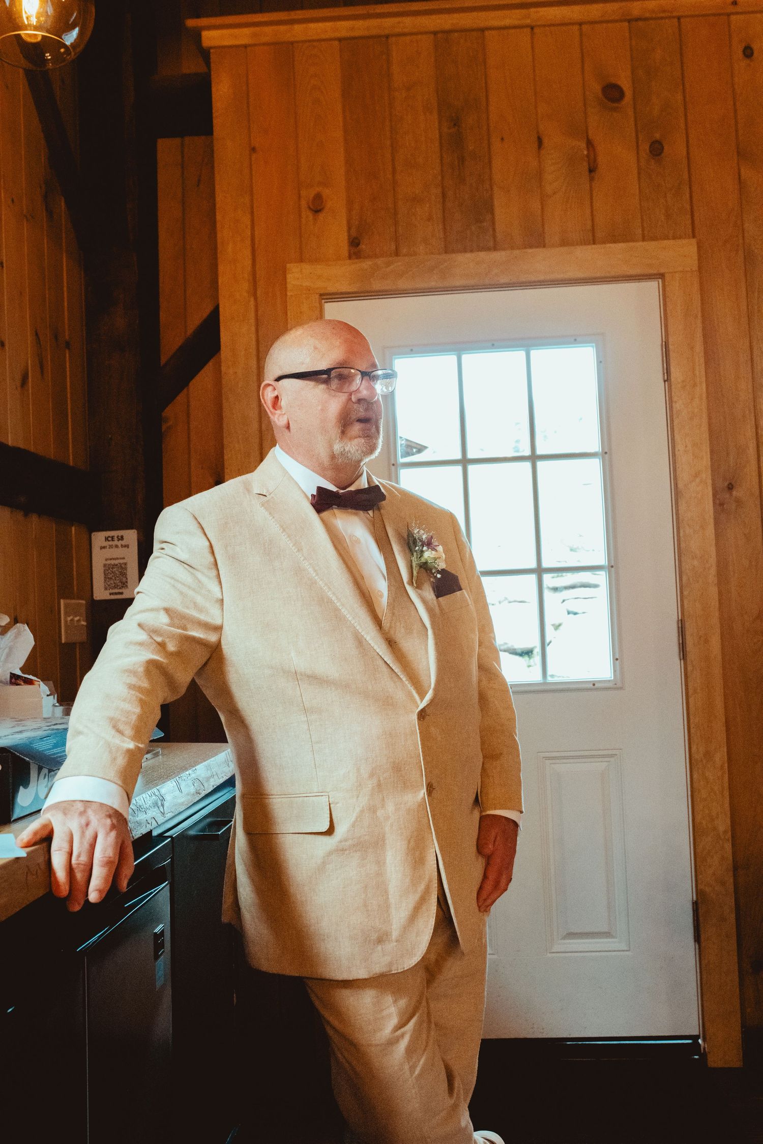 Bald groom in tan suit and bow tie with boutonniere stands in rustic wooden barn venue near white door.