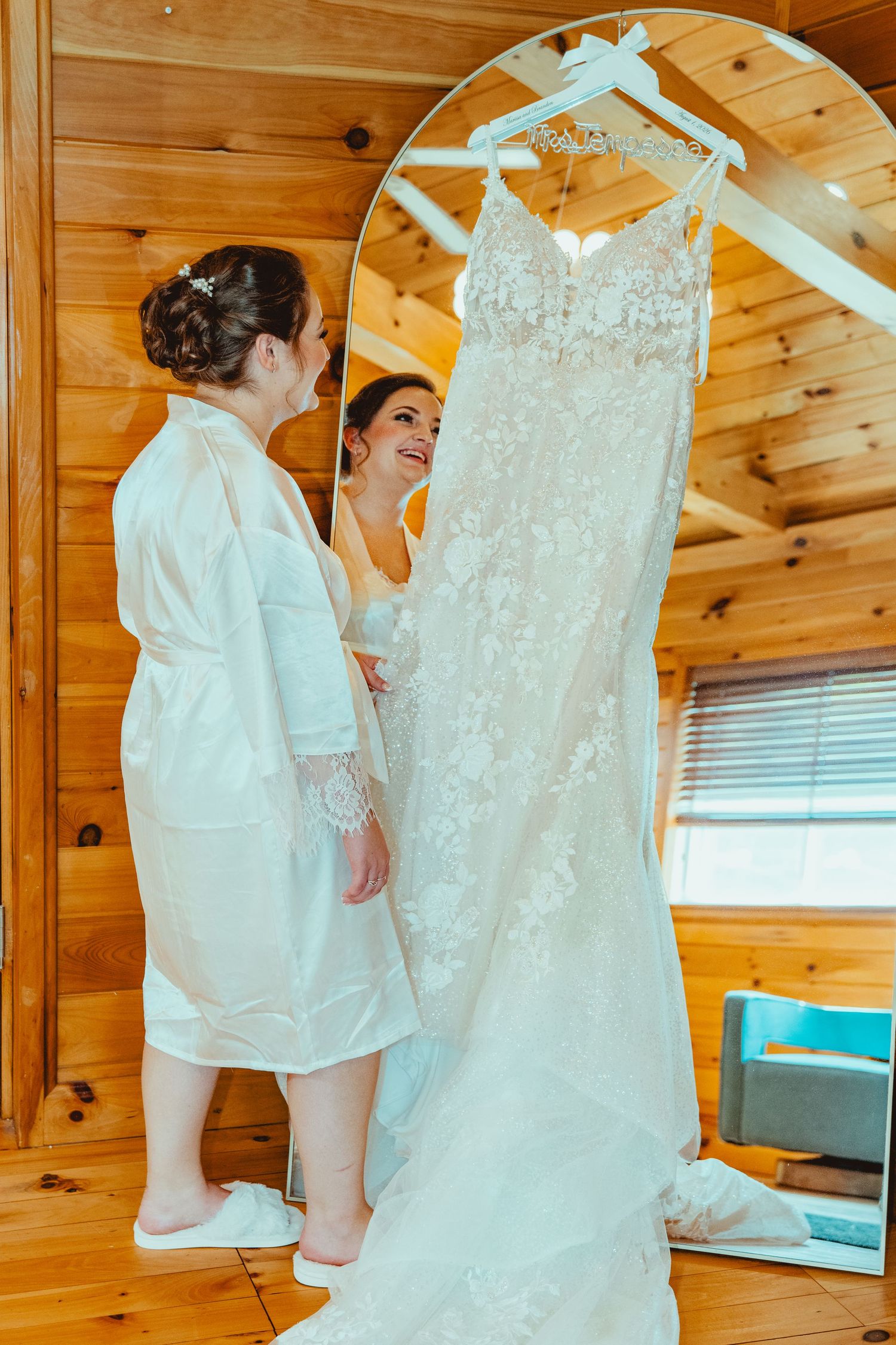 Two brides in white robes smiling beside a lace wedding dress hanging on a mirror in a cozy wooden cabin.