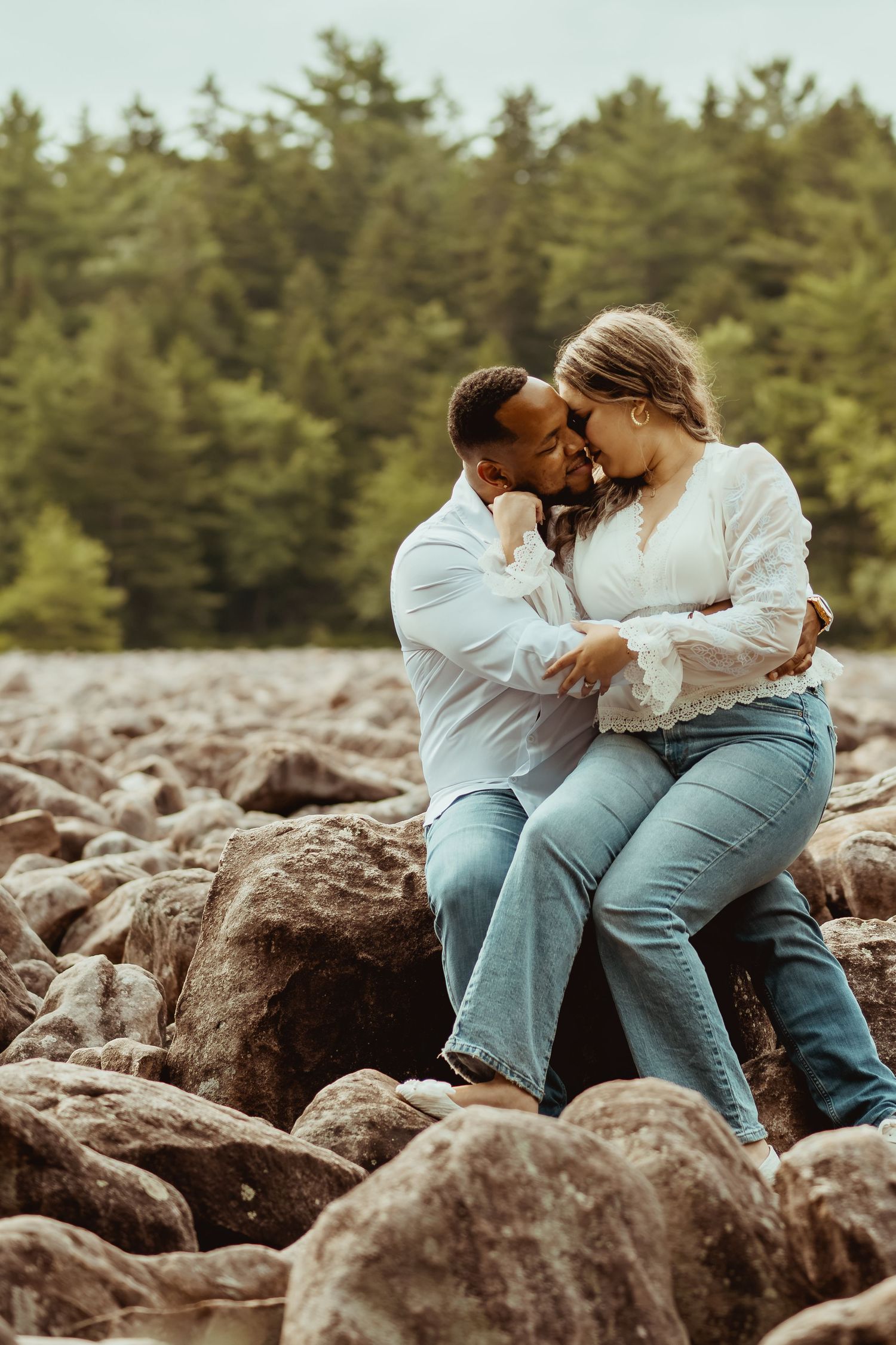 Romantic couple sitting on rocks embracing and kissing with evergreen forest backdrop.