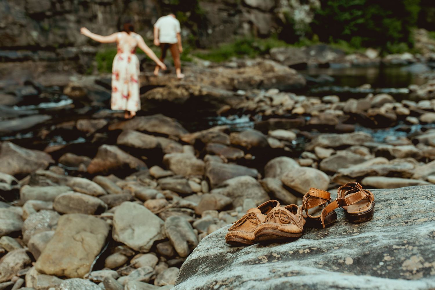 Sandals on rocky riverbank with couple holding hands in background during outdoor engagement session.