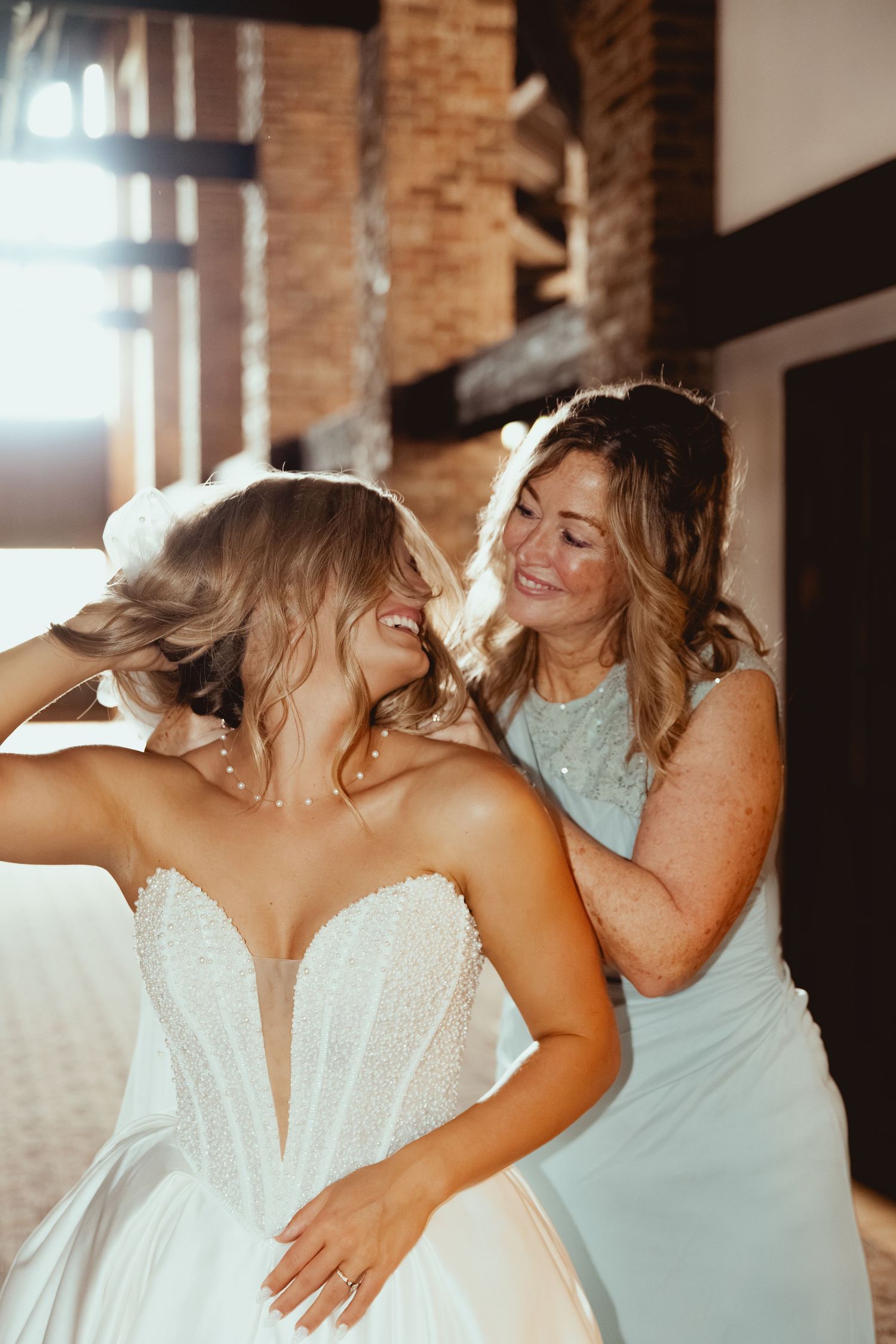 Bride smiling as bridesmaid helps adjust her veil in a warmly lit venue before the wedding ceremony.