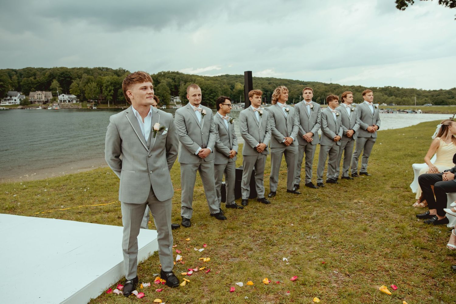 Groomsmen in gray suits lined up outdoors by a lake during a wedding ceremony on a cloudy day.