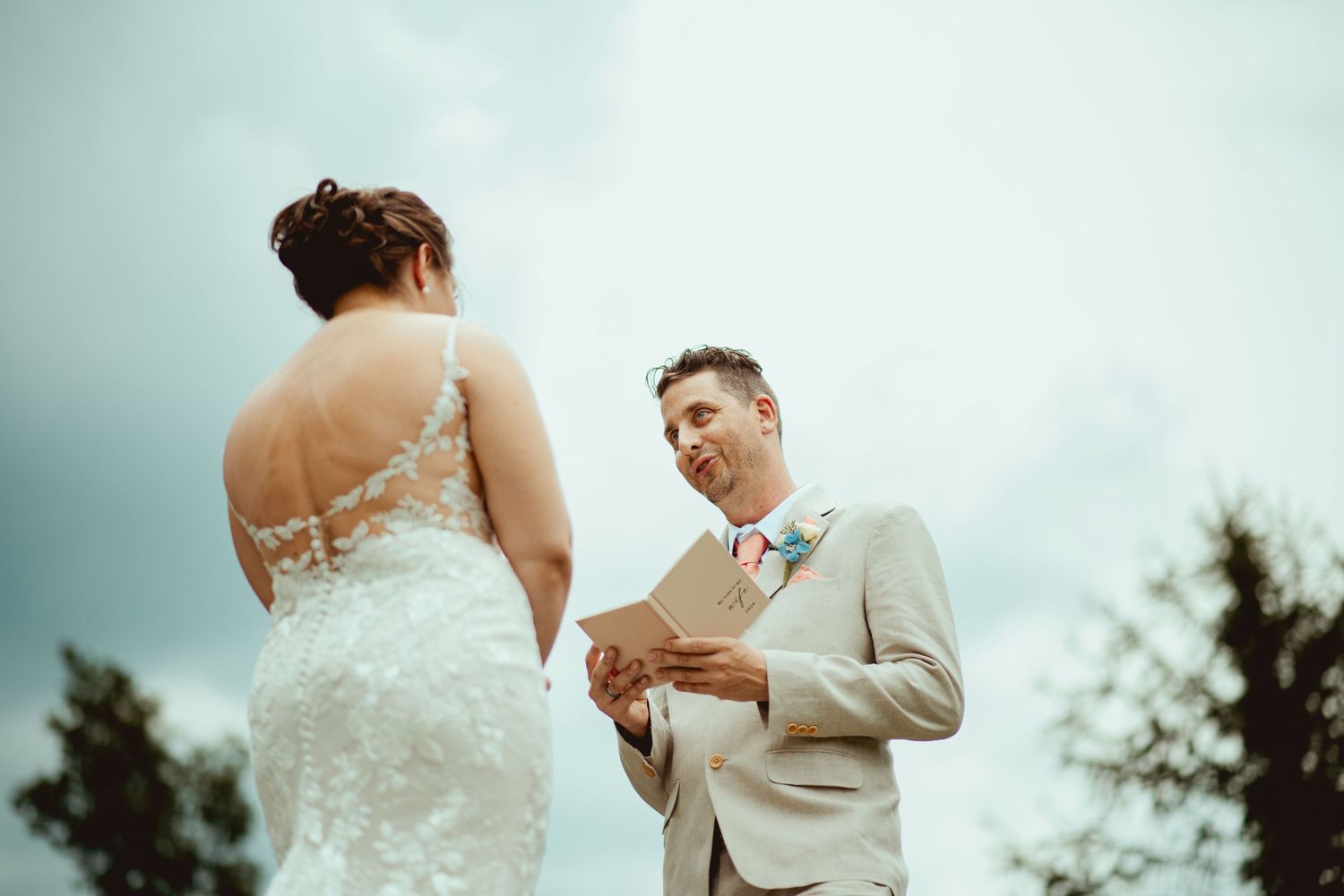 Groom in beige suit reads wedding vows from a card to bride in lace dress during outdoor ceremony.