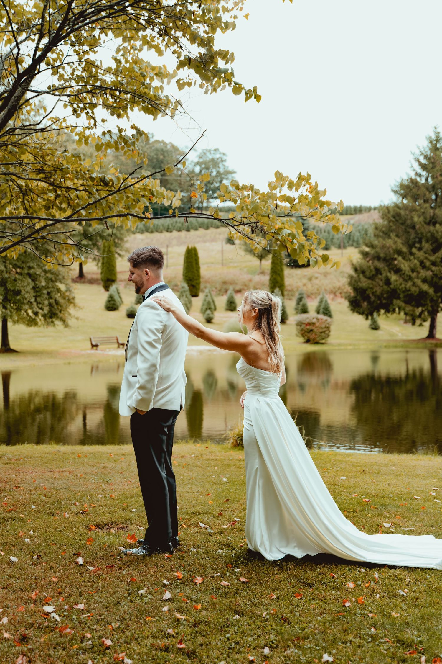 Bride in a white gown reaches to tap the groom's shoulder in a first look on an autumn lawn beside a calm pond.