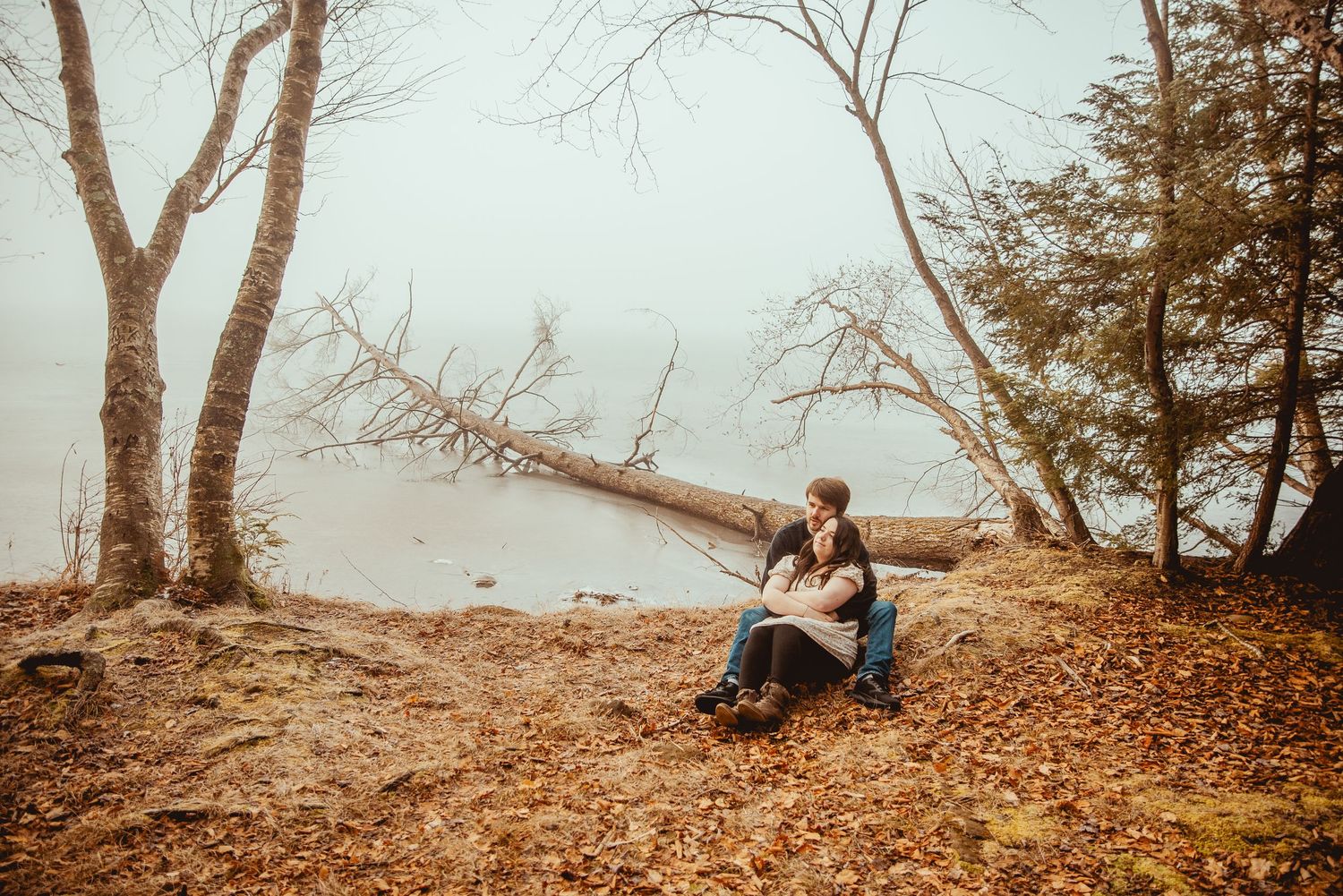 A couple sits together on rocks by a misty lake shore during autumn.
