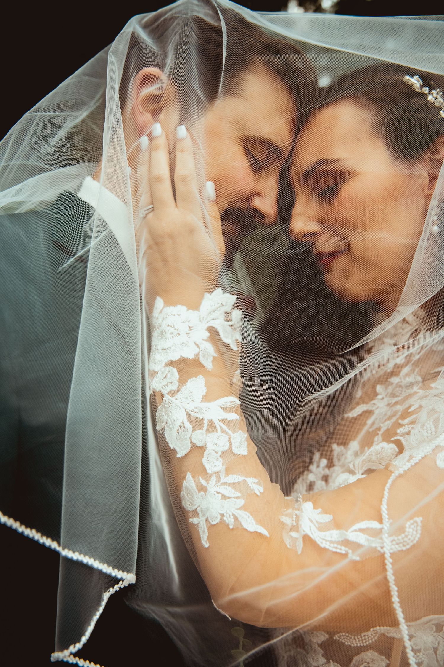 Bride and groom share intimate moment under wedding veil, bride wearing lace dress and tiara.