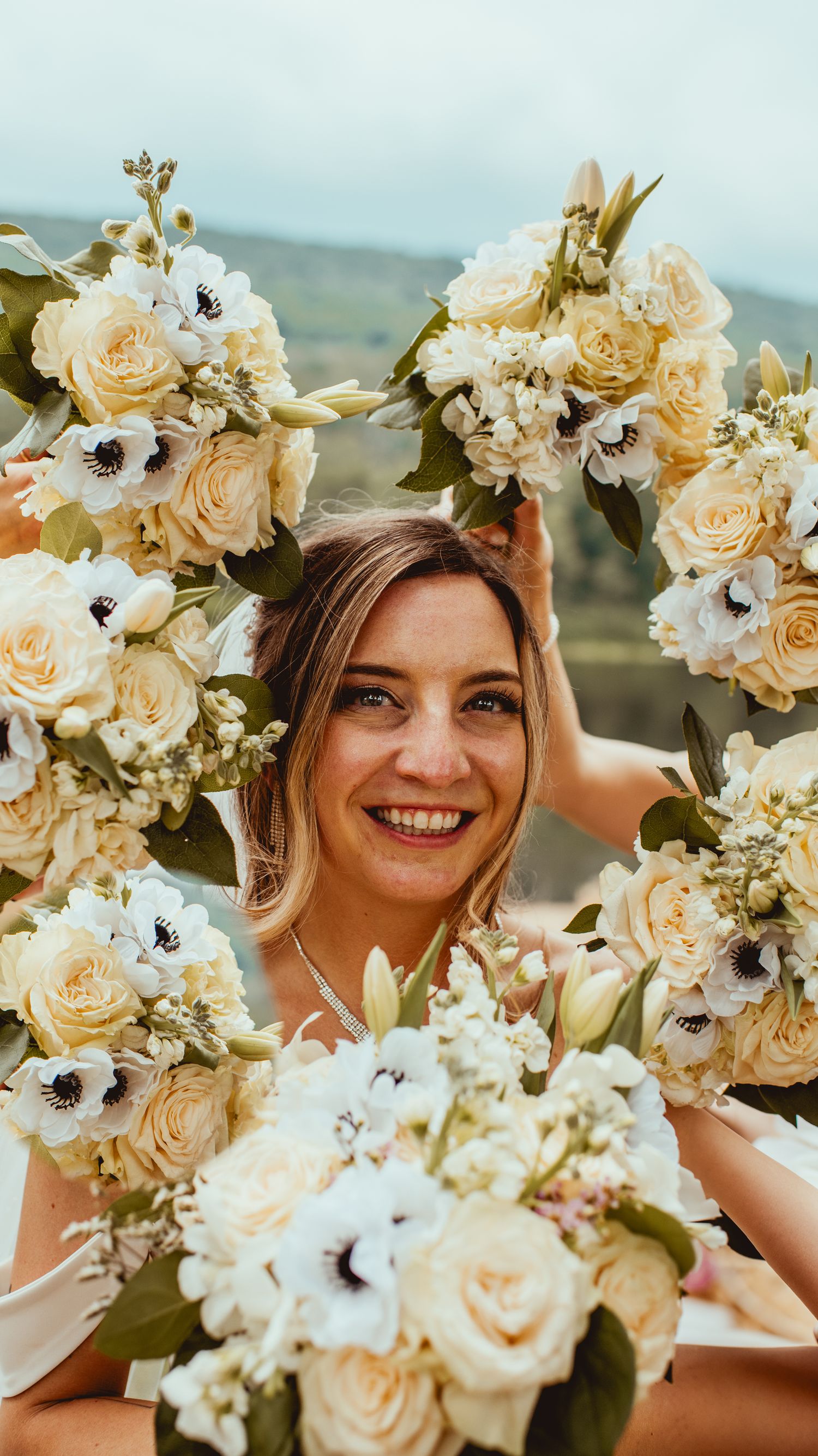 A floral crown made of peach roses and white anemones creates a romantic bridal look against a scenic outdoor backdrop.