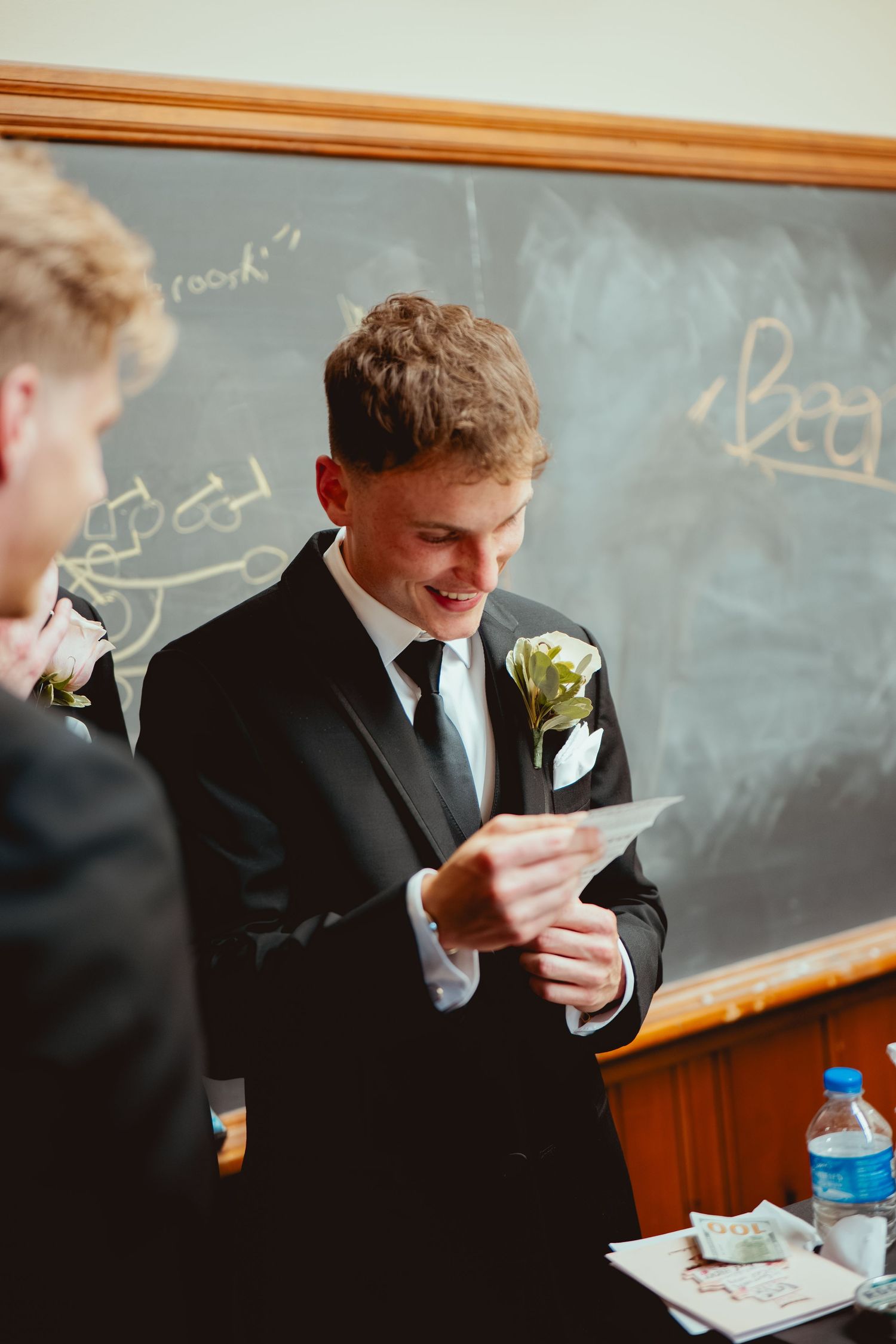 Groomsmen in black suits check their phones while standing near a chalkboard in a classroom setting.