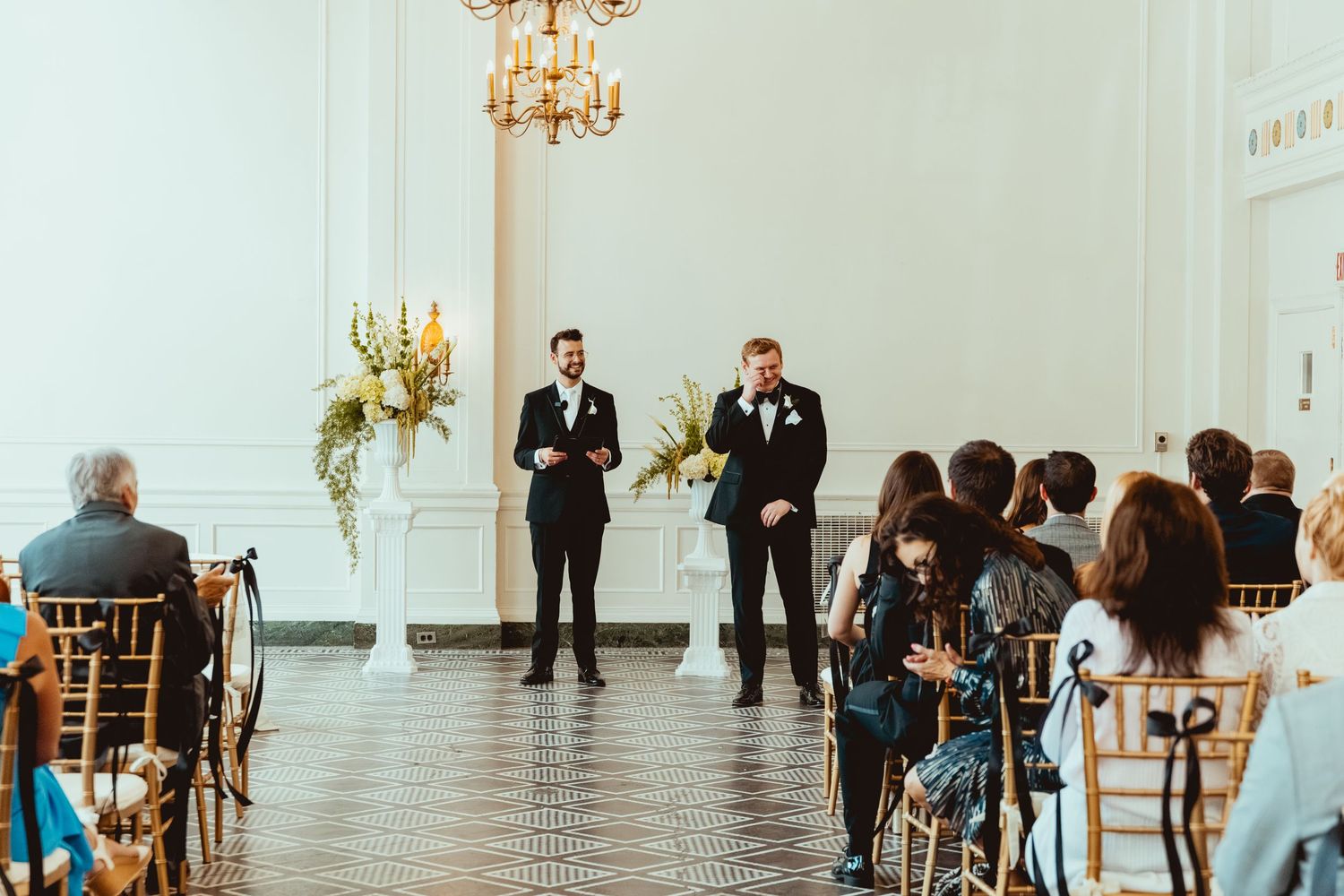 Elegant wedding ceremony in progress with gold chairs and chandeliers in a bright white ballroom.