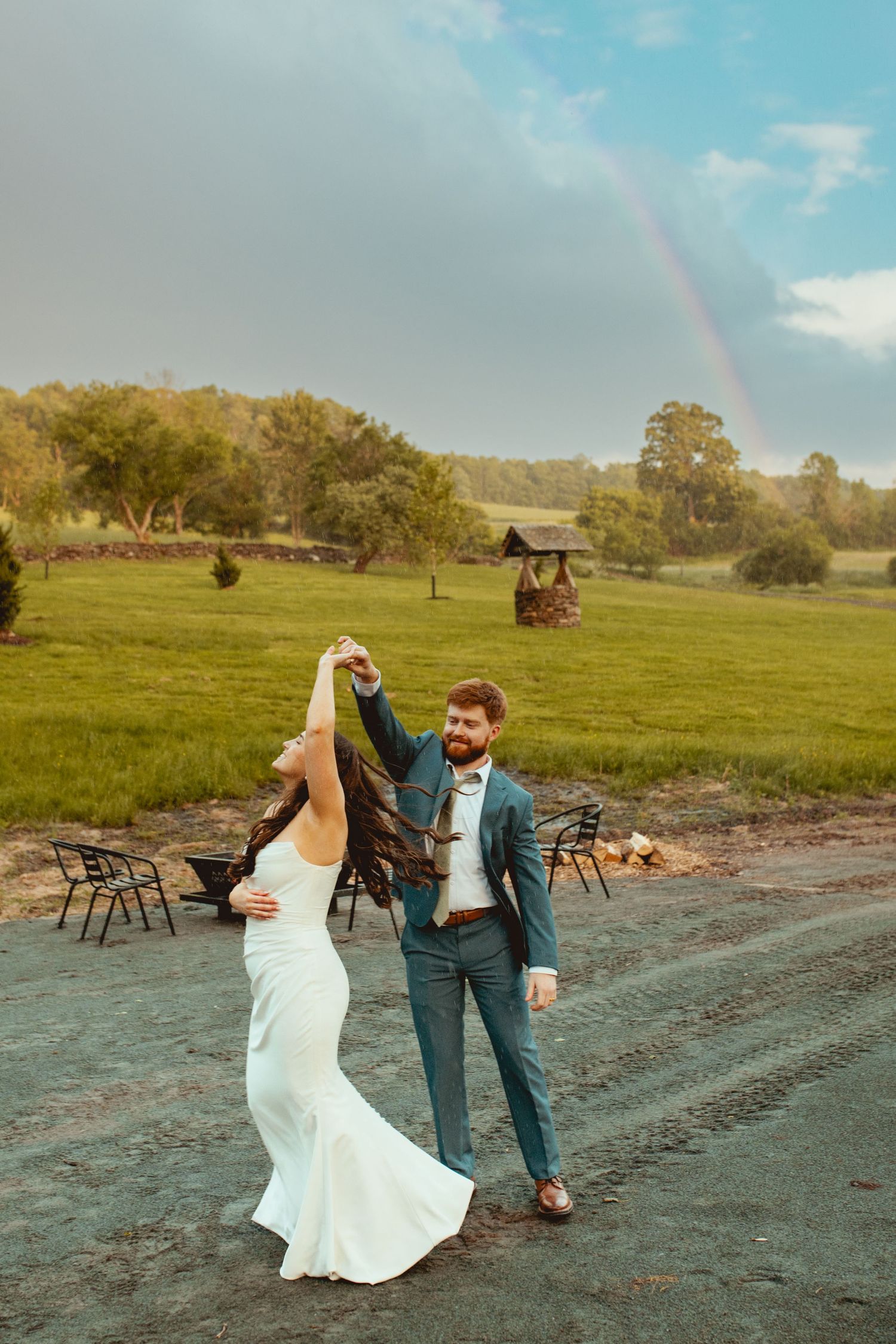 A couple dances joyfully on a dirt path with a rainbow and rustic barn in the countryside background.