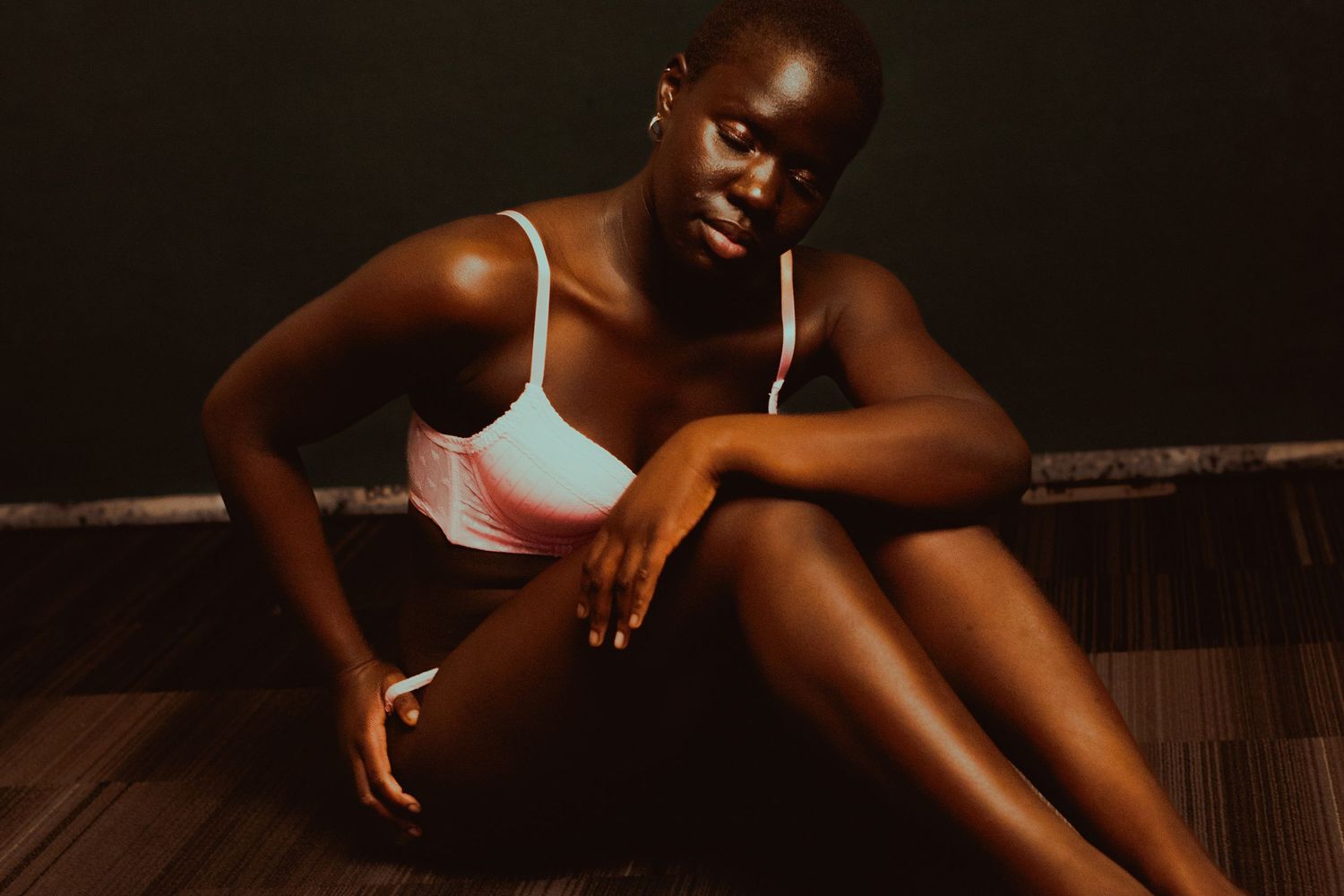 Young Black woman in a white sports bra sitting relaxed on a wooden floor against a dark background.