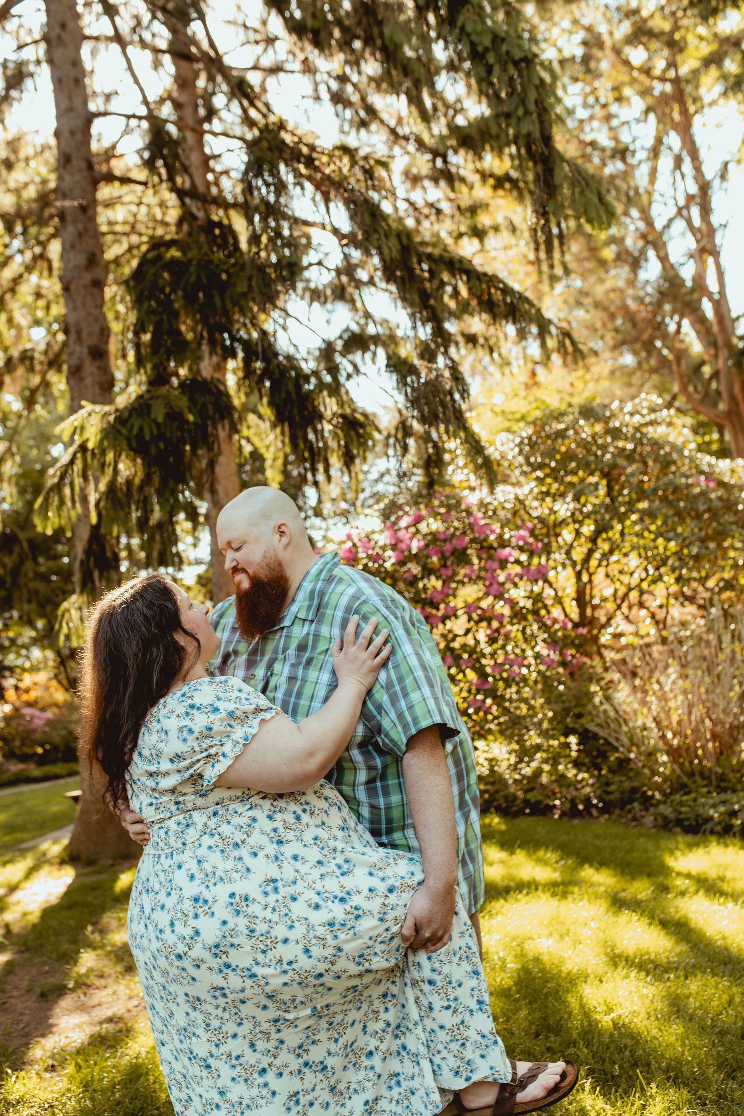 A couple shares a romantic kiss in a lush garden surrounded by blooming flowers and tall trees on a sunny day.
