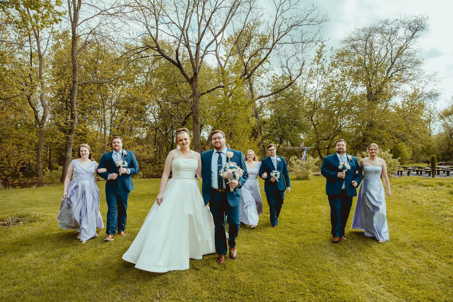 Wedding party walking through green park with bride in white gown and attendants in blue and grey.
