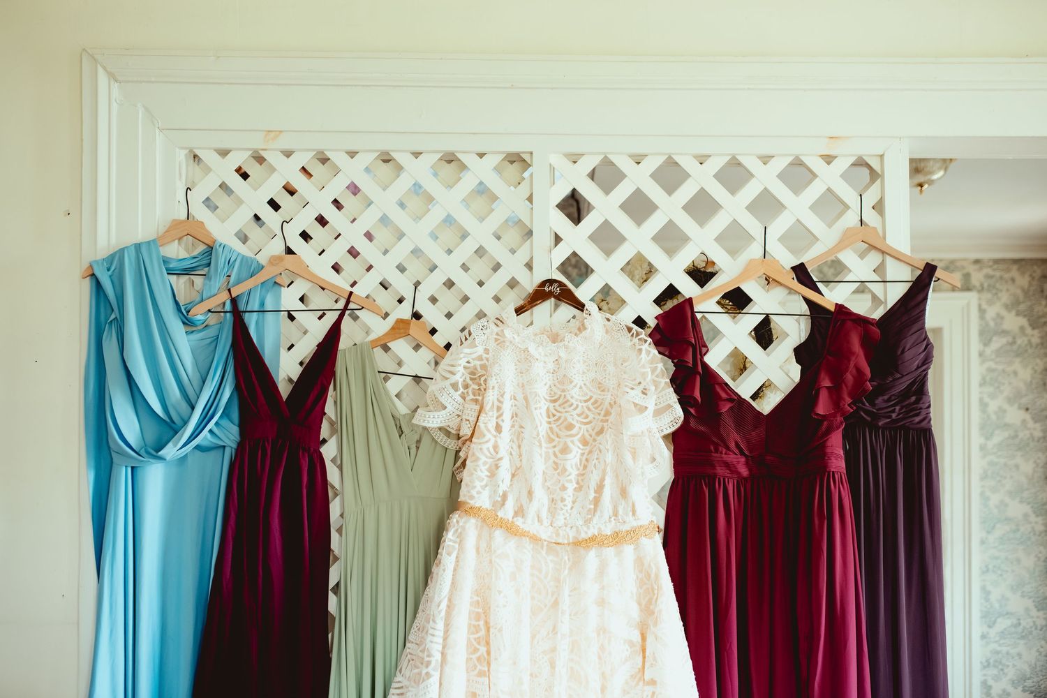 Burgundy and turquoise bridesmaid dresses hanging on white lattice backdrop with floral decorations.