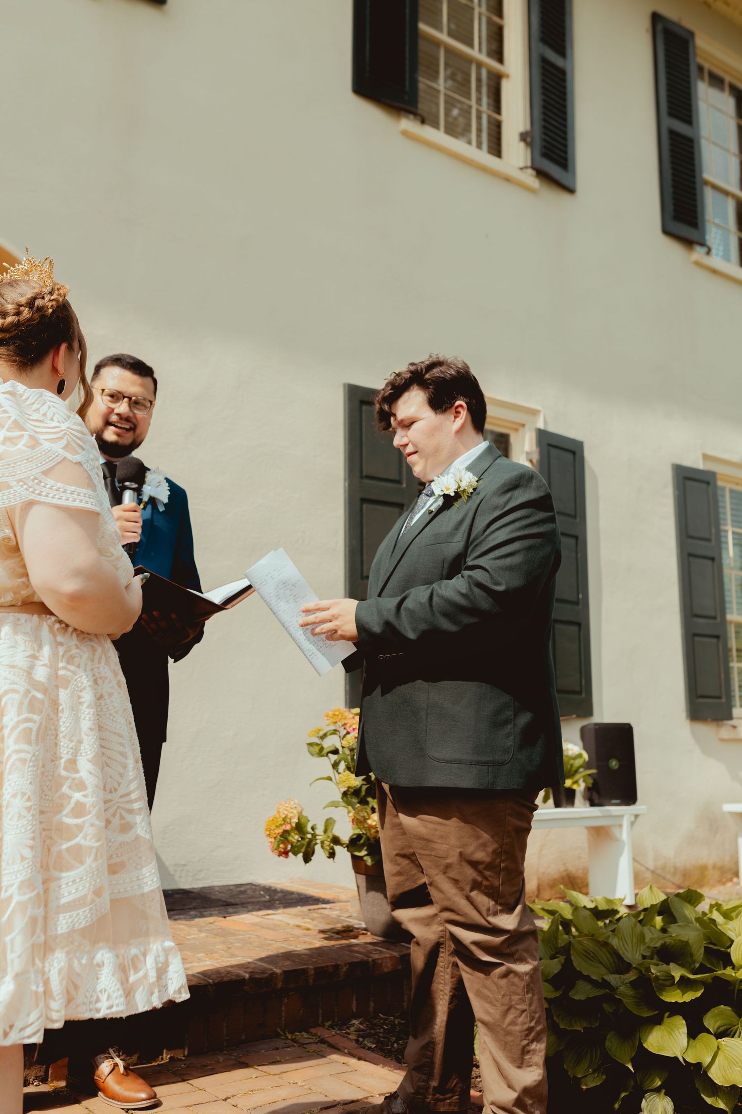 A couple reads their vows during an intimate outdoor wedding ceremony against a white building with shutters.
