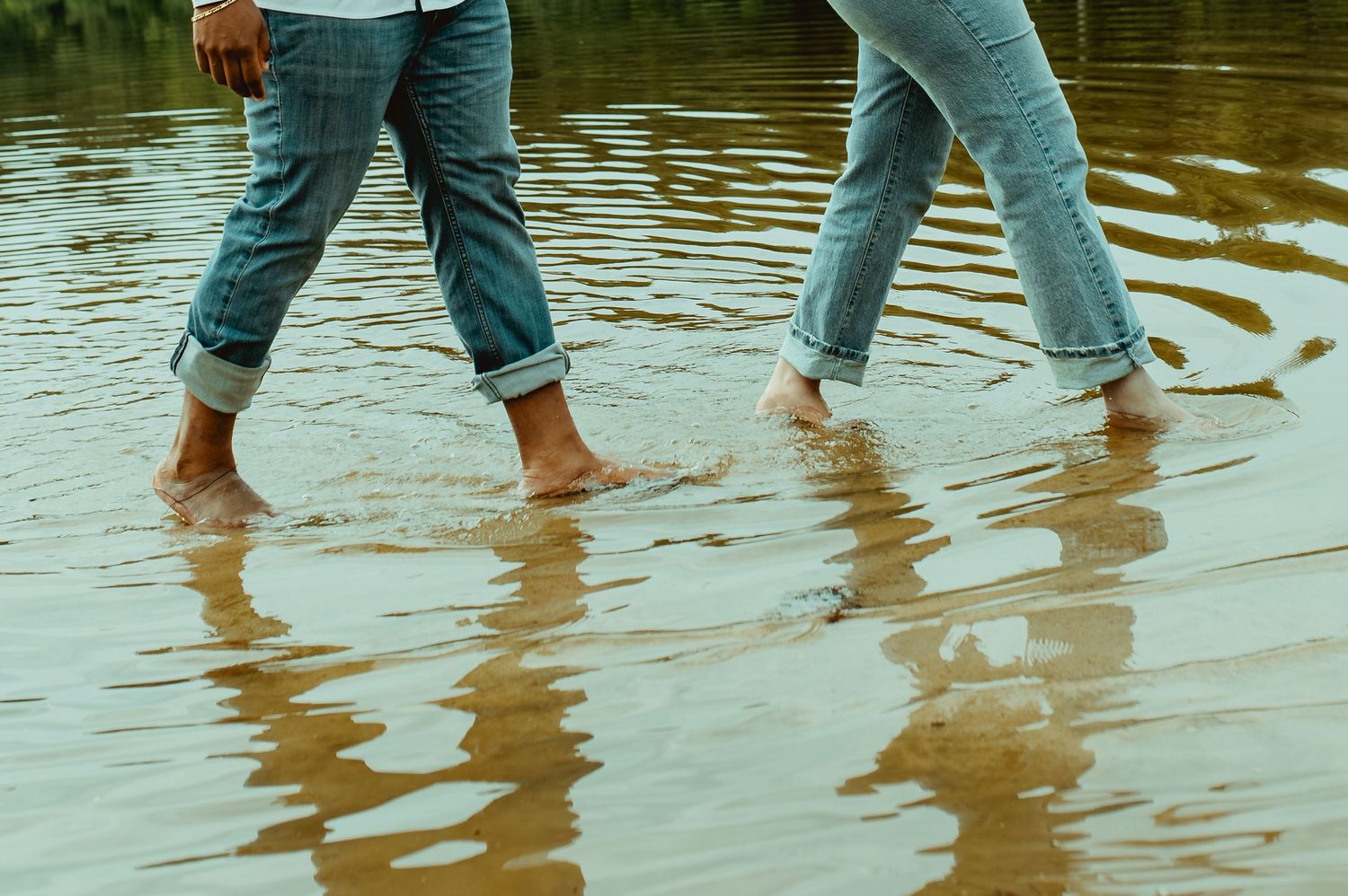 Two people walk barefoot through shallow water, wearing rolled-up jeans on a reflective sandy shore.
