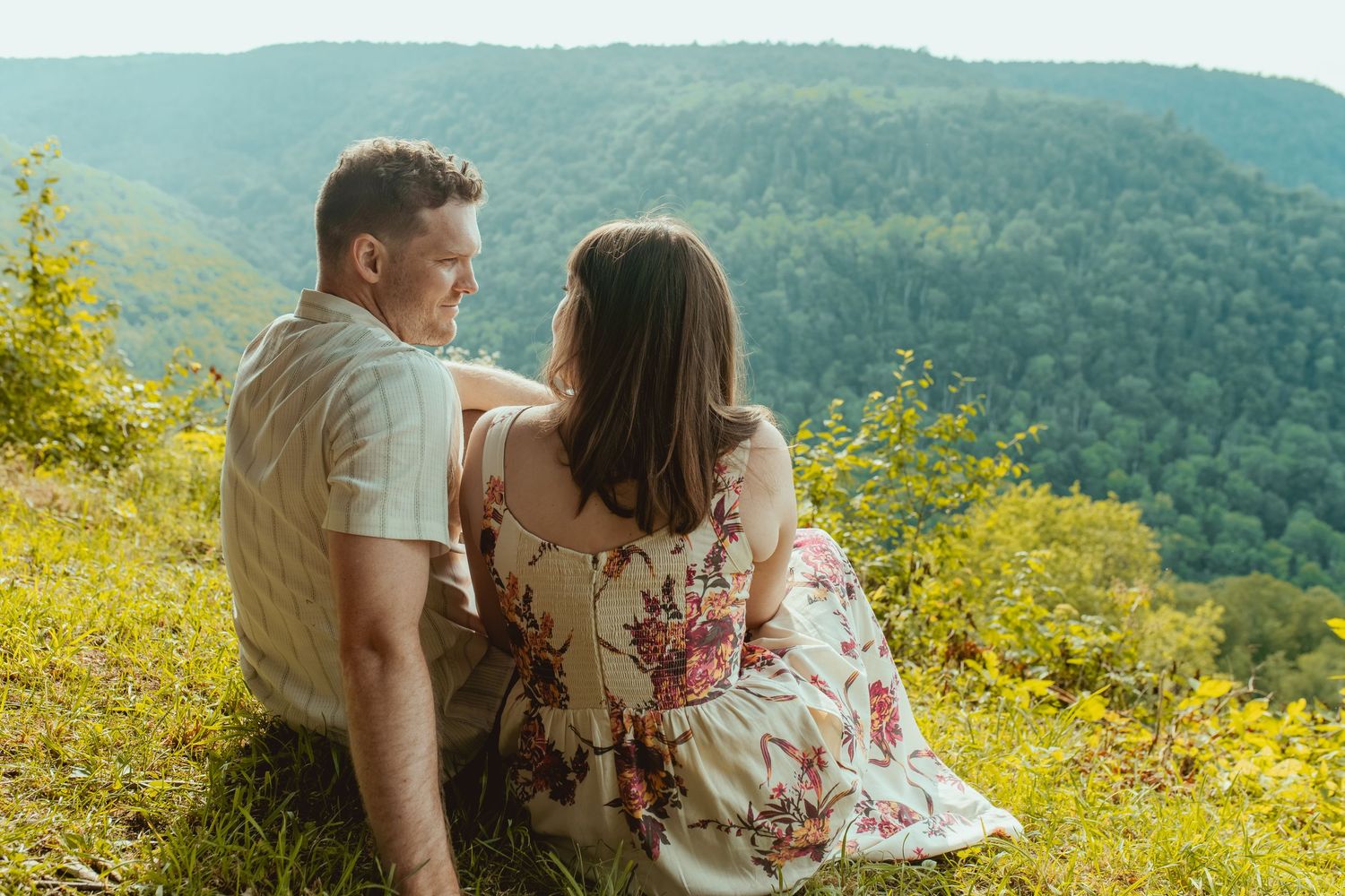 Couple sitting on a grassy overlook enjoying a scenic mountain valley view, woman in floral dress.