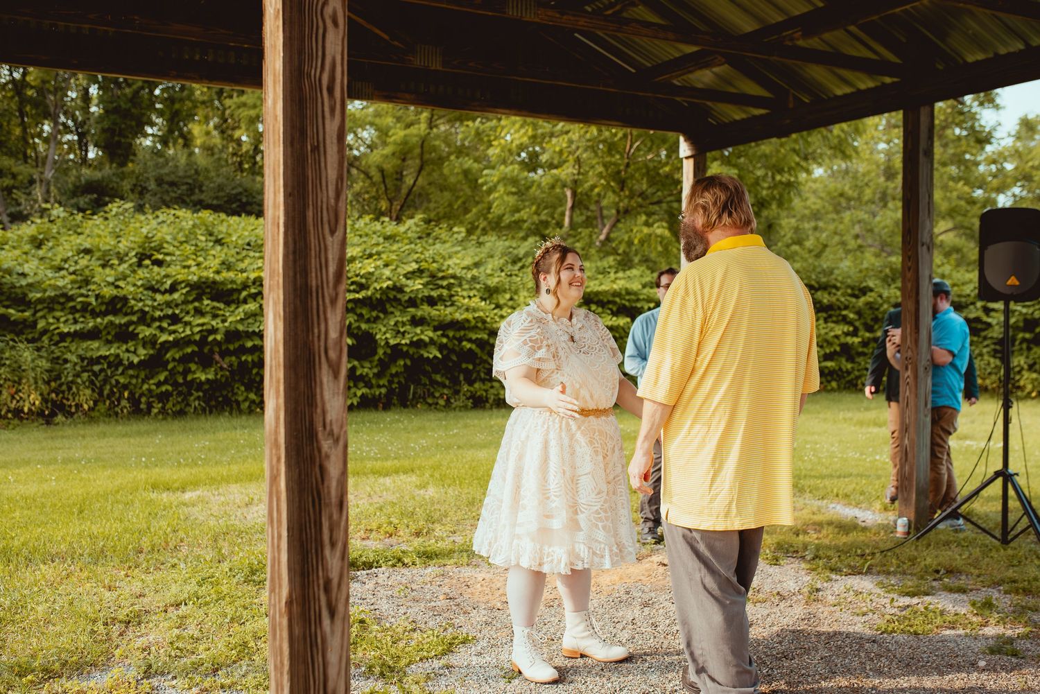 Two people in formal attire stand under a wooden pavilion during an outdoor celebration.