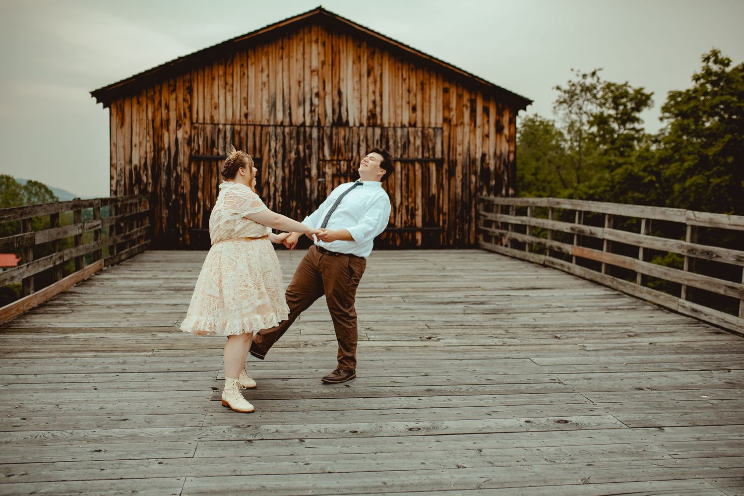 Couple standing on a rustic wooden boardwalk with weathered barn buildings in the background.
