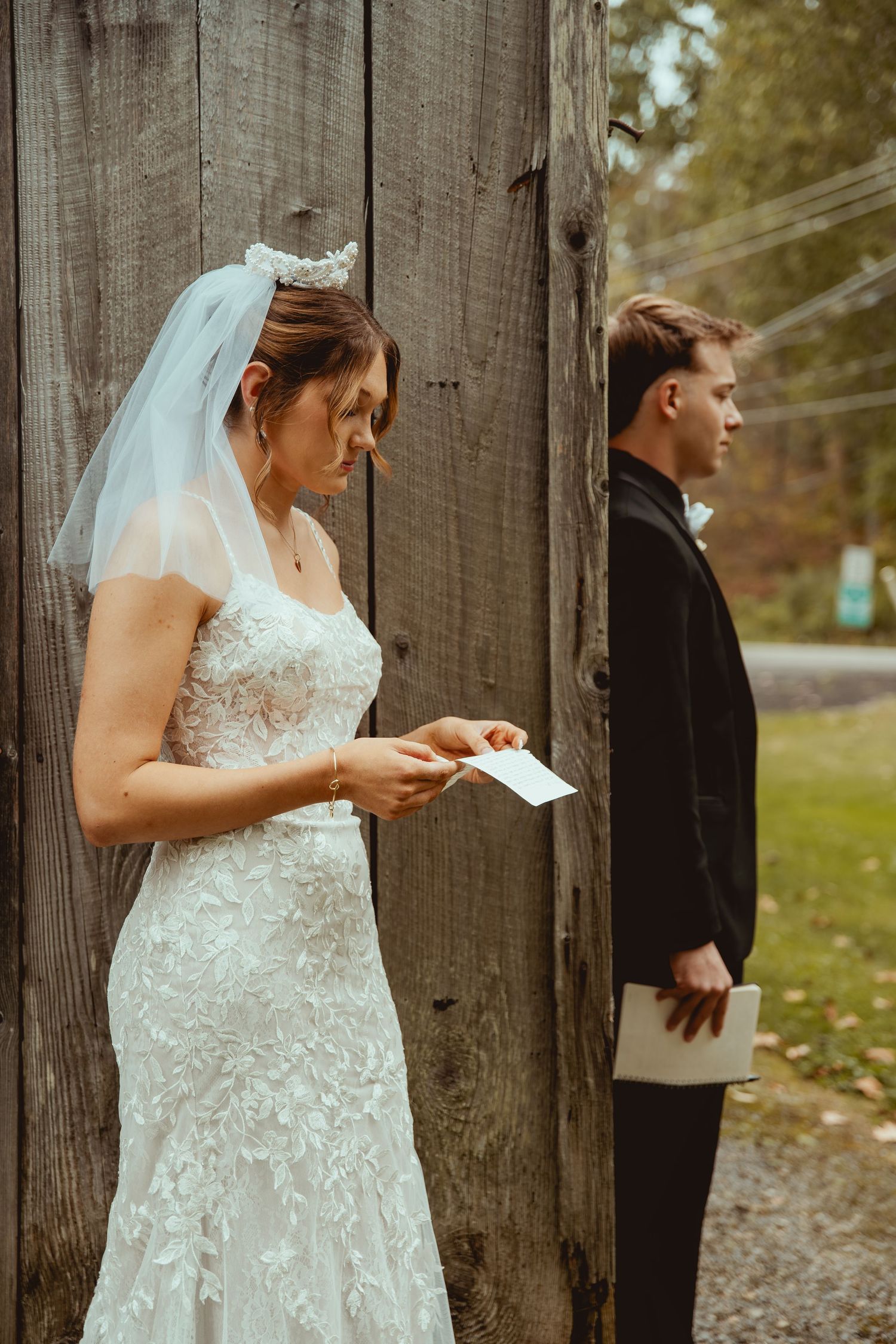 A couple exchanges letters while standing on opposite sides of a rustic wooden wall during their first look wedding moment.