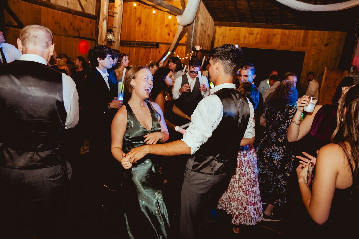 Wedding guests dance and celebrate on a dimly lit wooden dance floor at a rustic reception venue.