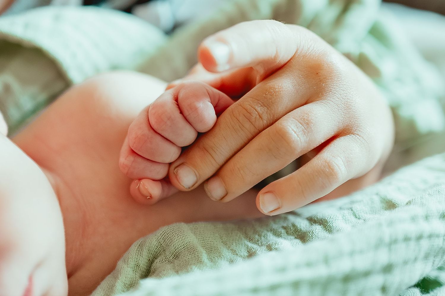 A tiny baby hand gently grasps an adult finger while resting on soft white fabric.